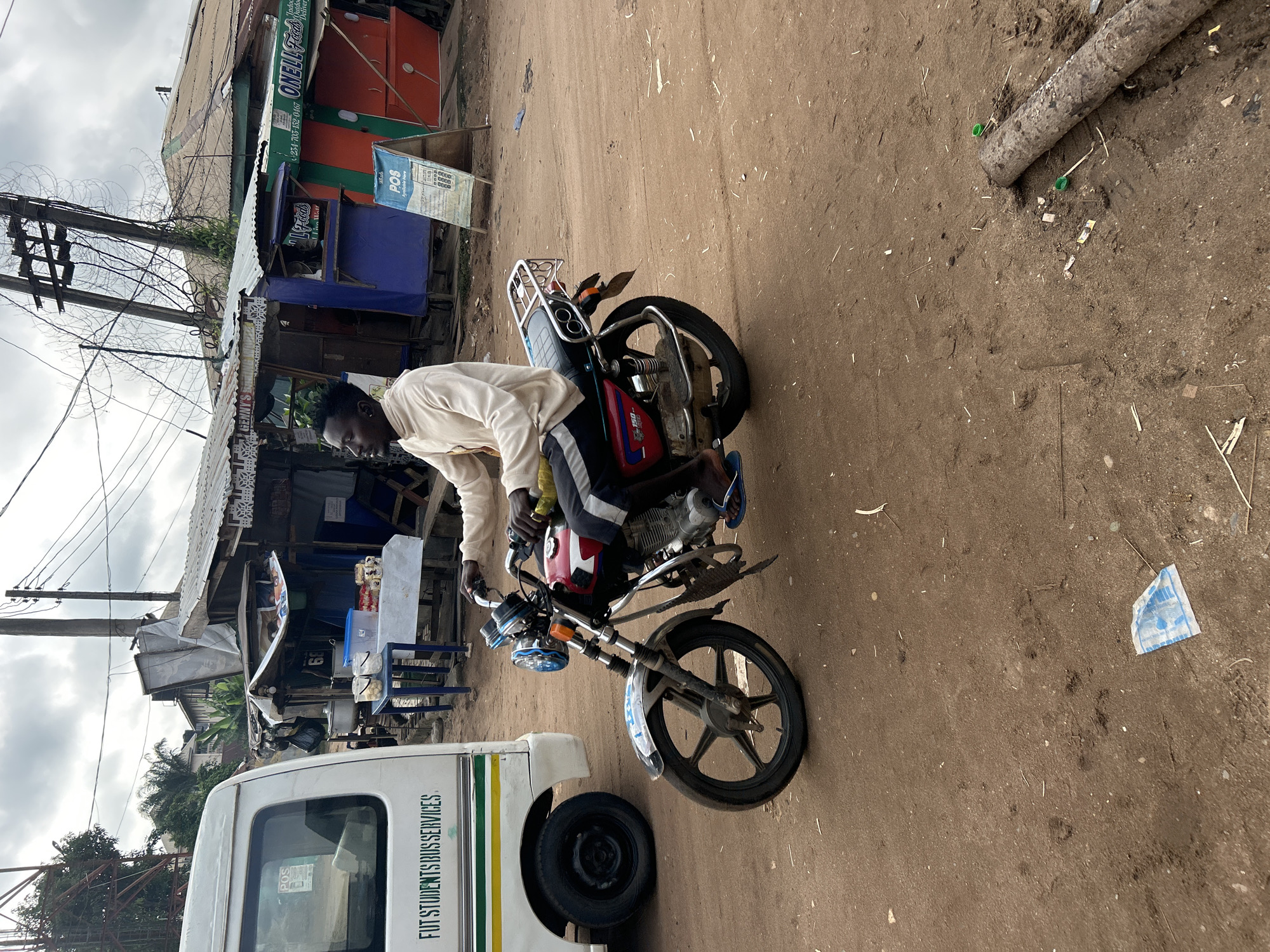 In Ihiagwa, Nigeria, on a dusty, unpaved road, a man is riding a red and black motorcycle. He is wearing a light-colored long-sleeved shirt and dark pants, with blue flip-flops on his feet. His right hand is on the handlebars, and his left arm is resting on the bike. The motorcycle has a chrome rack on the back and blue accents on the headlight. To the left of the motorcycle is a white bus with green lettering that reads "FUT STUDENTS BUS SERVICES." In the background, there are several makeshift market stalls with corrugated metal roofs. One stall has a sign for "ONELL FOODS." Overhead, power lines crisscross the sky, and a cloudy sky suggests it may be overcast. The ground is a mix of dirt and scattered debris, including a piece of blue and white patterned paper and a log with two green bottle caps on it.