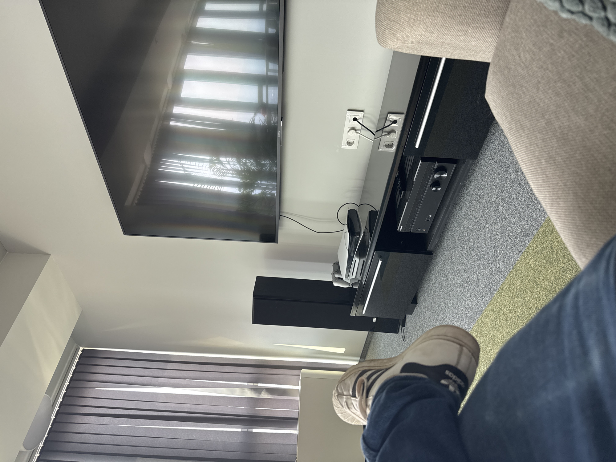 A first-person perspective shot shows a person's legs and feet in the foreground, with their right foot crossed over their left leg. The person is wearing blue jeans and white Adidas sneakers. In the background, a modern living room is visible. A large, flat-screen television is mounted on the wall above a black entertainment console. The console holds several electronic devices, including a sound system and a gaming console. A tall black speaker stands to the left of the console, and vertical blinds cover a large window behind it, allowing natural light to filter into the room. The flooring consists of grey carpet with a striped green and yellow runner rug. The overall ambiance suggests a contemporary, well-equipped living space, possibly located in a modern apartment building within the city of Sofia, Bulgaria, given the architectural style and interior design elements.
