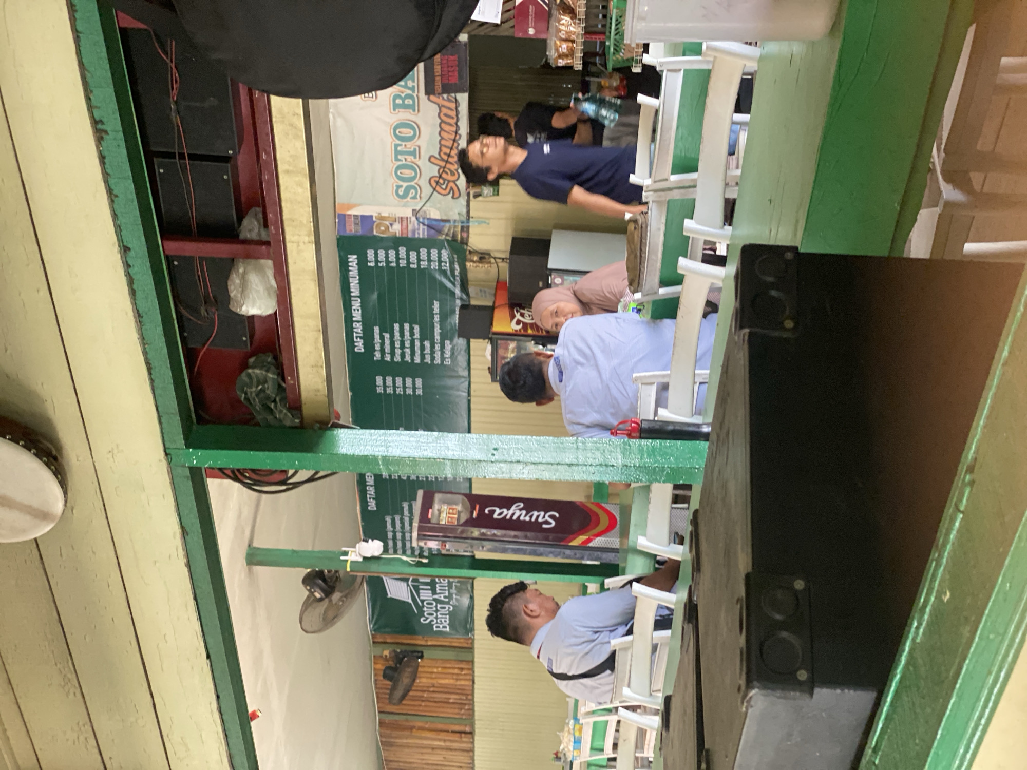 The image depicts an indoor seating area of a restaurant, likely in Banjarmasin, Indonesia, identified by a prominent sign for "Soto Bang Amang". The scene is casual, with diners seated at green-painted tables and white chairs. In the foreground, a black, rectangular object with visible circular fixtures sits on a green surface, possibly a counter or table. A ceiling fan is visible above and to the left.
In the mid-ground, several people are seated. On the left, a man in a light blue shirt is looking down. To his right, a woman in a pink hijab is seated at a table. Further to the right, a man in a dark blue shirt is walking away from the camera, carrying a bottle of water. Behind him, another person in a dark shirt is visible.
The background reveals more seating and a wall adorned with a bamboo-like texture. A menu board is mounted on the wall, displaying various food and drink items with prices, including "DAFTAR MENU MINUMAN" (Drink Menu List) and specific items like "Teh es/panas" (Iced/Hot Tea) and "Sirup es/panas" (Iced/Hot Syrup). Another sign, "SOTO BANG AMANG" with "SELAMAT" written below it, suggests the establishment's name and a welcoming message. There are also shelves with packaged goods visible on the right side of the image. The overall atmosphere suggests a local eatery.