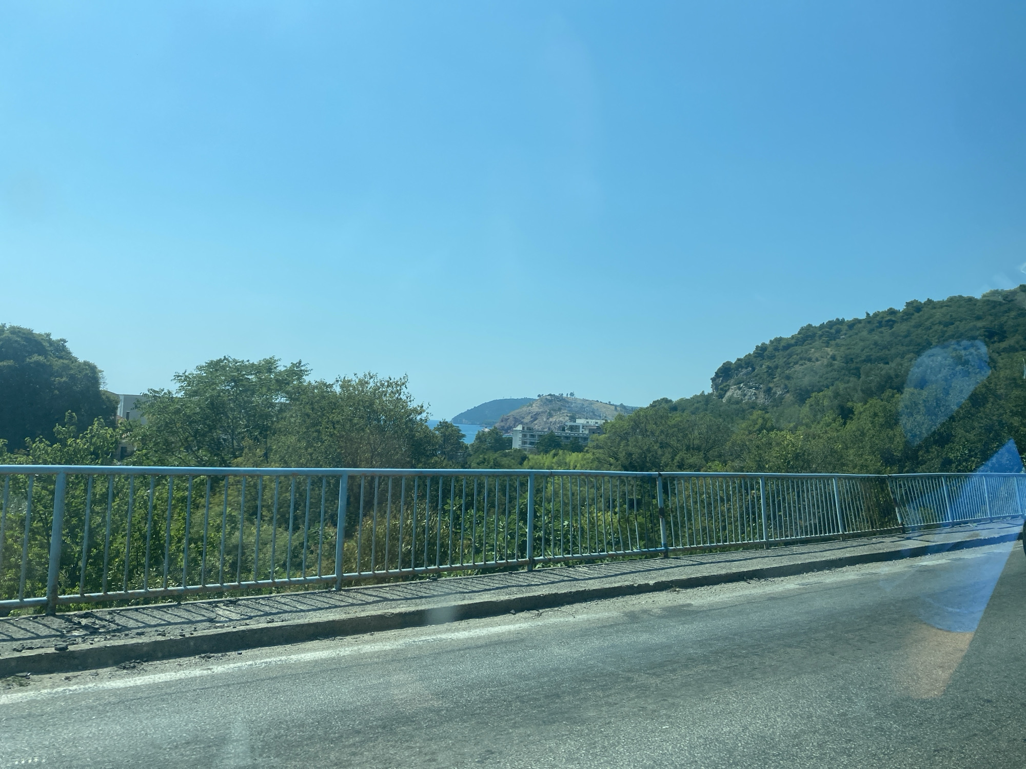 The image captures a view from a bridge, looking towards a coastal landscape. The foreground features a worn asphalt road and a light blue metal railing. Beyond the railing, lush green trees and foliage are visible, with some buildings peeking through the dense greenery. In the distance, a hill with structures and a body of water can be seen under a clear blue sky. The scene suggests a sunny day in a Mediterranean setting. Specifically, this appears to be a bridge within the municipality of Bar, overlooking the town of Sutomore, Montenegro.