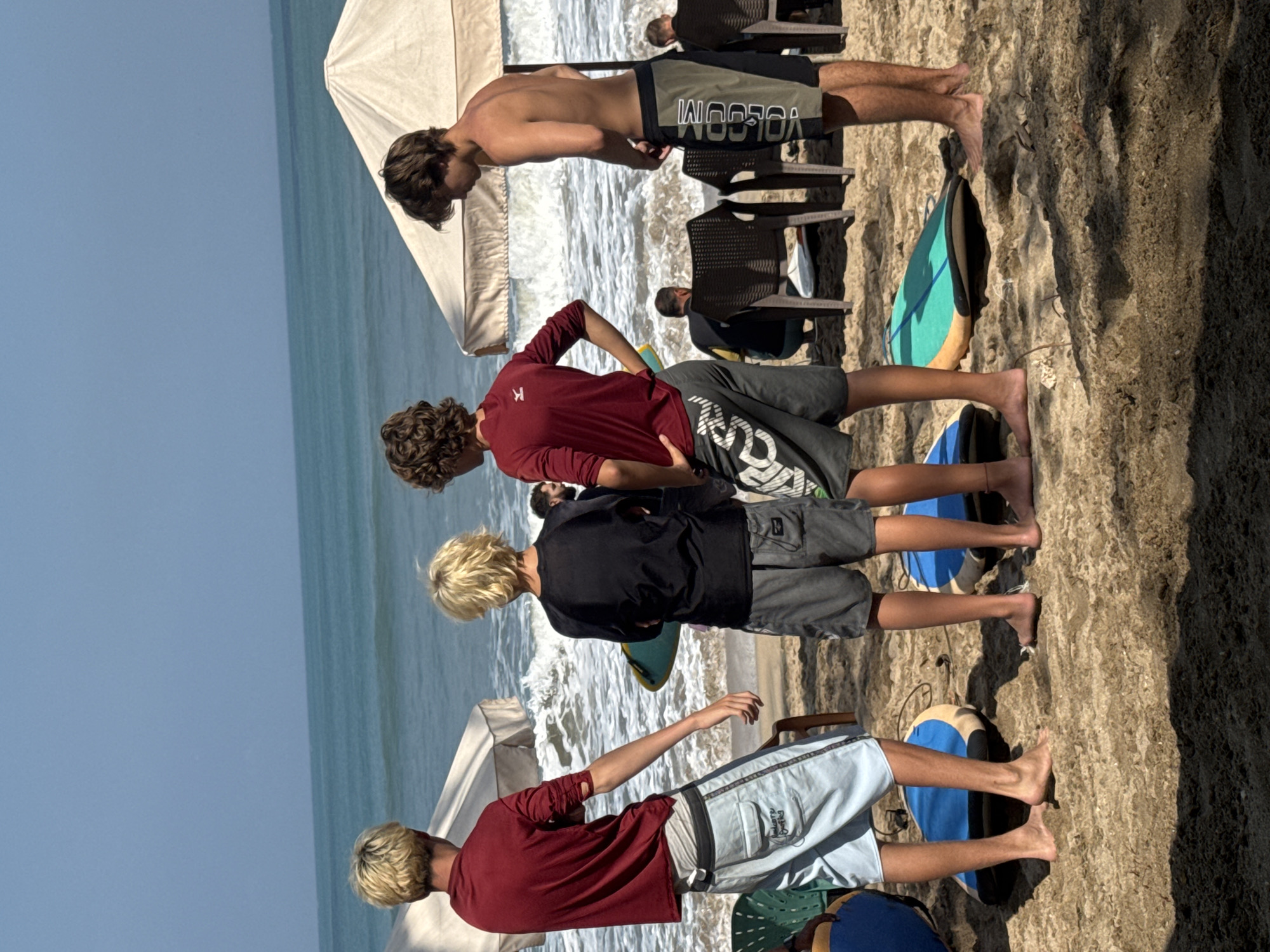 In Kuta, Indonesia, four young men stand on a sandy beach with the ocean and gentle waves in the background. Two men in the foreground, facing away from the camera, wear maroon long-sleeved shirts and light blue shorts, and black long-sleeved shirts and grey shorts respectively. Between them lie two blue and yellow surfboards. Another young man, shirtless, faces partially away from the camera, wearing dark grey and olive green swim trunks. Further back, another young man in a maroon shirt and grey shorts is visible. Two white umbrellas provide shade on the beach, and several plastic chairs are set up behind the surfboards. The scene appears to be a surfing lesson or a group preparing to surf.