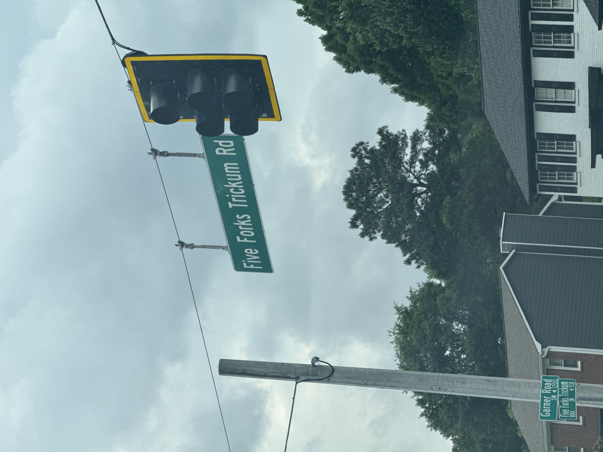 The image shows an intersection in Mountain Park, United States of America, marked by street signs and a traffic light. One street sign reads "Five Forks Trickum Rd". Another sign below it shows "Garner Road SW 1200" and "Five Forks Trickum Road SW 5330". The sky is overcast, suggesting cloudy weather. Trees and rooftops of buildings are also visible in the background.