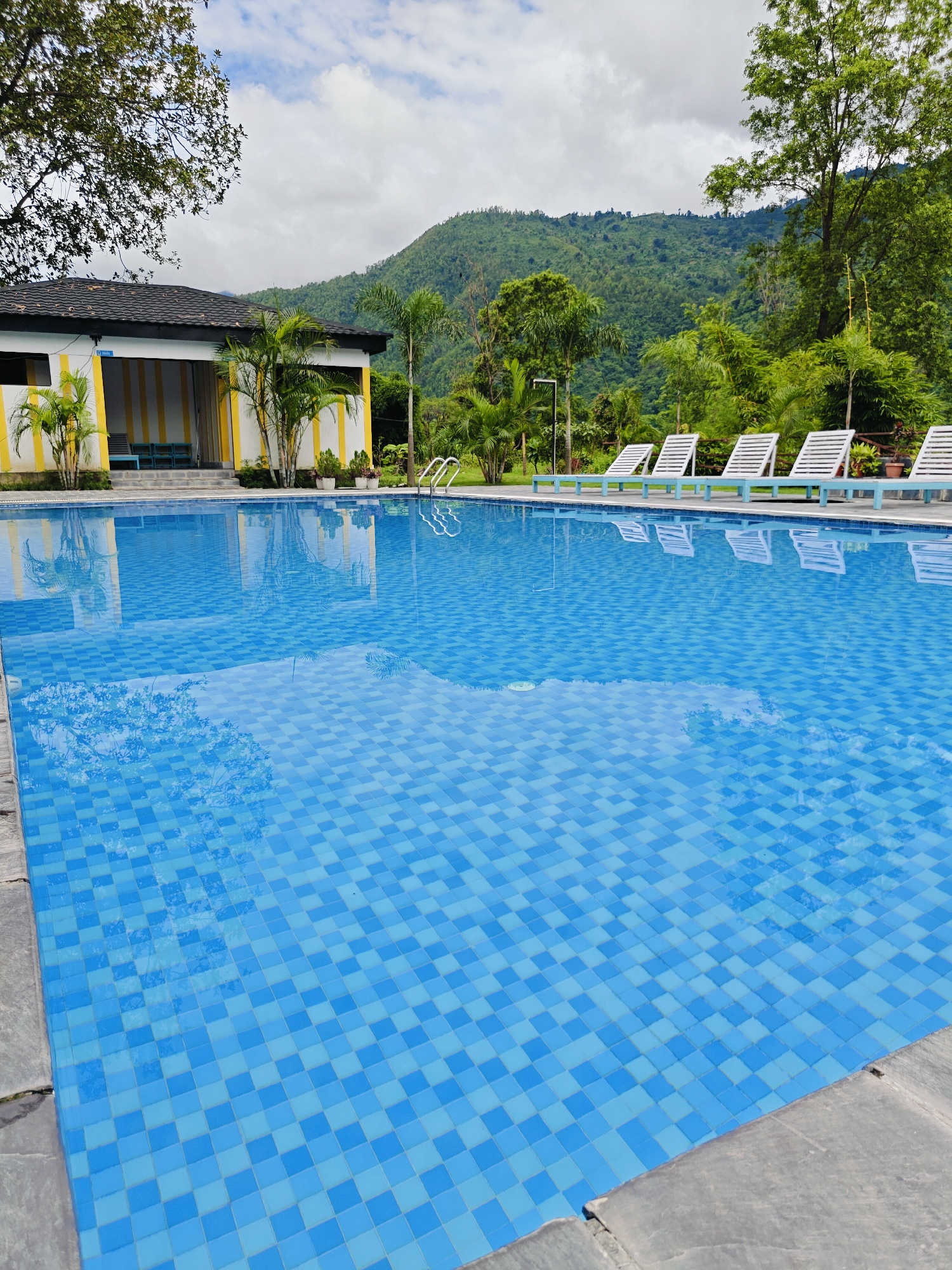 The image shows a large, clear blue swimming pool with a checkered tile pattern. In the background, there is a white building with yellow accents, surrounded by palm trees and lush green hills. Several white lounge chairs are lined up beside the pool. The sky is partly cloudy, and the overall scene depicts a serene resort or hotel setting, possibly located in Ichchhakamana, Nepal, known for its natural beauty.