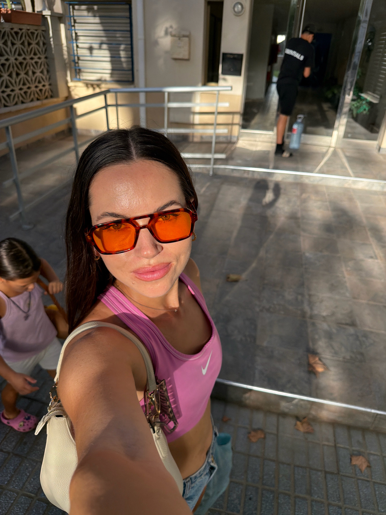 A woman in her late 20s or early 30s takes a selfie with a young girl behind her. The woman is wearing a pink sports bra and denim shorts, with orange-tinted square sunglasses and a white shoulder bag. Her hair is dark and long, and she is smiling at the camera. The young girl, who appears to be around 5-7 years old, is wearing a light purple tank top and shorts. She is facing away from the camera, looking down at something.
In the background, a man in a black t-shirt with white text and black shorts is walking into a building. He is holding a blue water bottle. The setting appears to be an outdoor walkway or street, with tiled ground and a building with large glass doors. The time of day seems to be late afternoon or early evening, as indicated by the warm, angled sunlight casting shadows. The presence of fallen leaves on the ground suggests it might be autumn.
Visible text includes "ESSENTIALS" on the back of the man's t-shirt and a white Nike logo on the woman's sports bra. The overall mood is casual and summery. The location is identified as Salou, Spain.