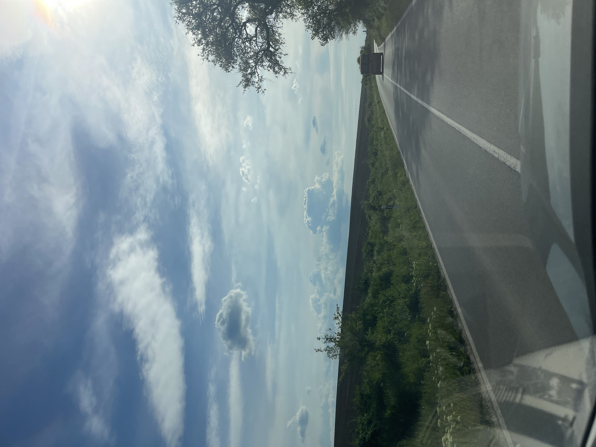 The image shows a view from inside a car driving on a road. The road has a white line marking the center and is bordered by green vegetation. A vehicle is visible ahead in the distance. Above, a clear sky with scattered clouds is observed. The presence of trees is discernible in the upper portion of the frame. Vetovo, Bulgaria cannot be confirmed as the location as there are no specific indicators to definitively connect the scene to that city.