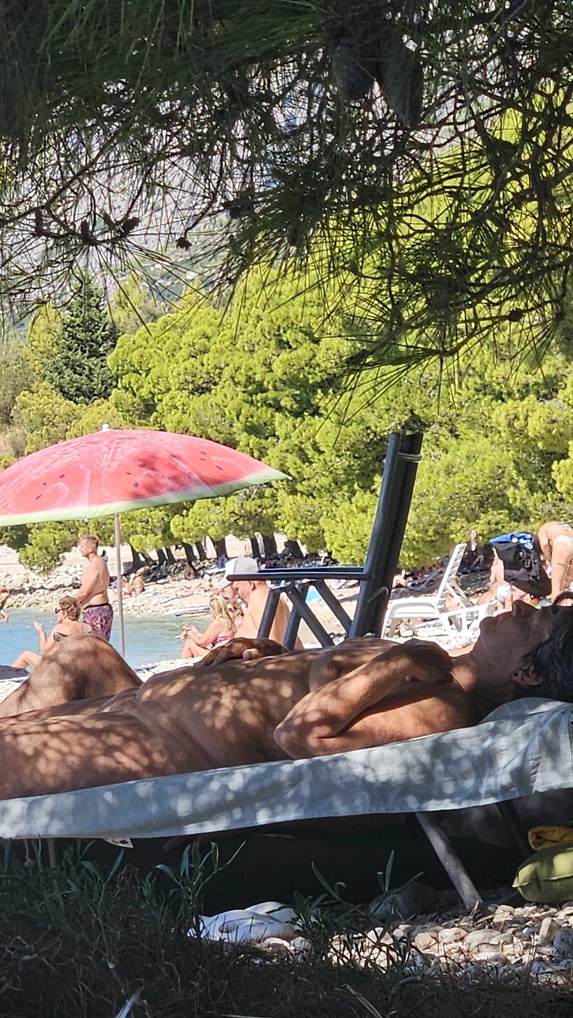 The image depicts a beach scene in Općina Podgora, Croatia, with several people visible. In the foreground, a person lies on a lounge chair, sunbathing. They are partially obscured by shadows from overhead foliage. Further back, a red and green watermelon-themed umbrella is set up on the pebble beach, providing shade for a group of people sitting nearby. Some individuals are visible in the background, engaged in beach activities. The sea is visible to the left, with a wooded hillside forming the backdrop. The overall atmosphere suggests a sunny day at the beach.