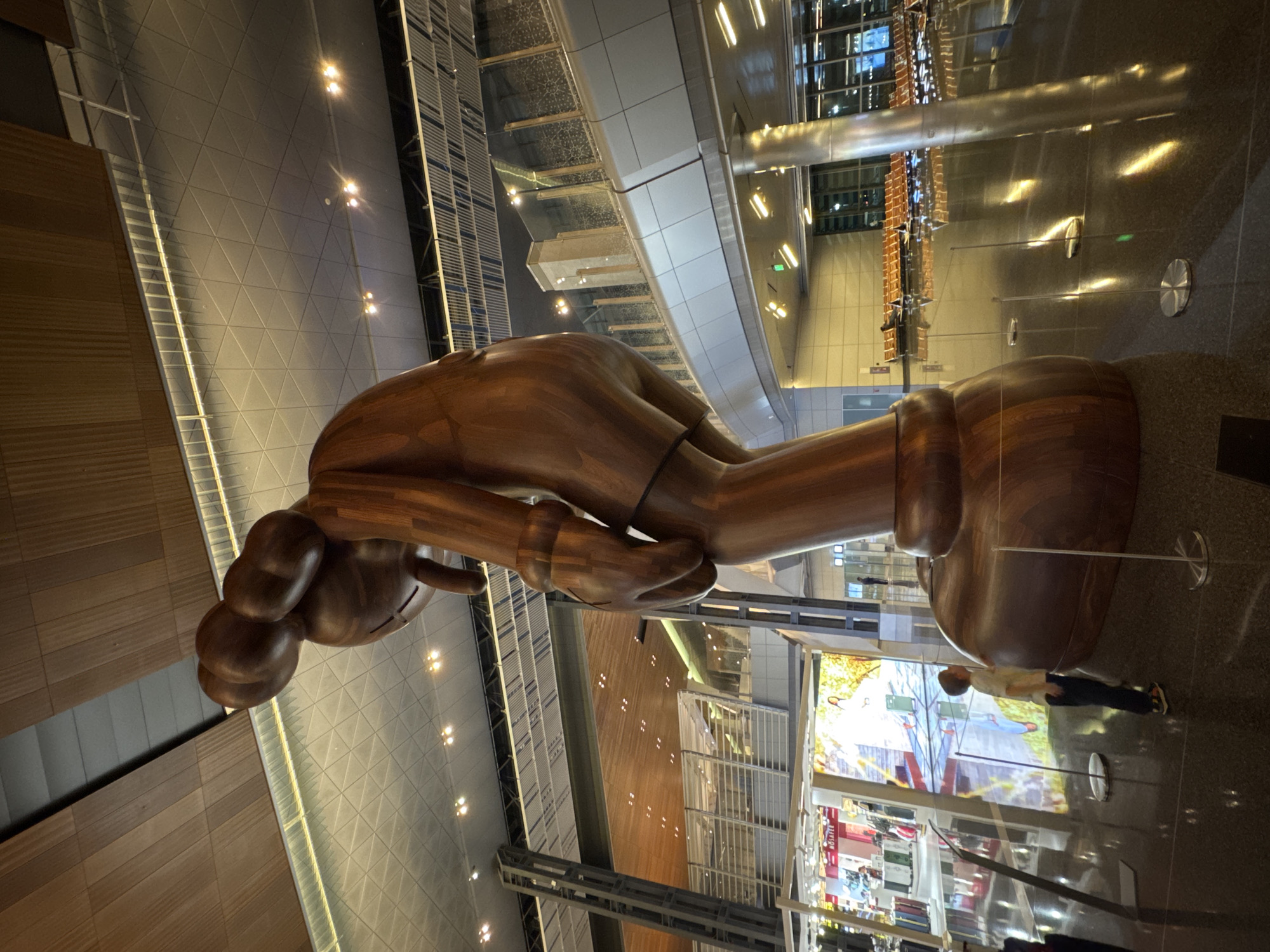 The image shows a large wooden sculpture of a character with a bent posture, in the center of an indoor space. The sculpture is made of polished wood and has smooth, rounded forms. In the foreground, a young boy in casual clothing stands to the left of the sculpture, looking up at it. The environment appears to be a modern, spacious building with a high ceiling and reflective floors. The ceiling is a complex structure of wood panels and metallic elements, with recessed lighting. In the background, there are rows of empty seats, suggesting a waiting area or a public concourse. There are also shopfronts and large digital screens displaying imagery. The architectural style and the presence of modern amenities suggest this is a public space, likely an airport terminal or a large exhibition hall. The image was taken in the Hamad International Airport in Doha, Qatar, which is known for its large-scale art installations. The lighting in the scene is artificial, casting reflections on the polished floor and highlighting the smooth surfaces of the sculpture. The overall impression is one of a contemporary, well-lit, and architecturally significant indoor public space.