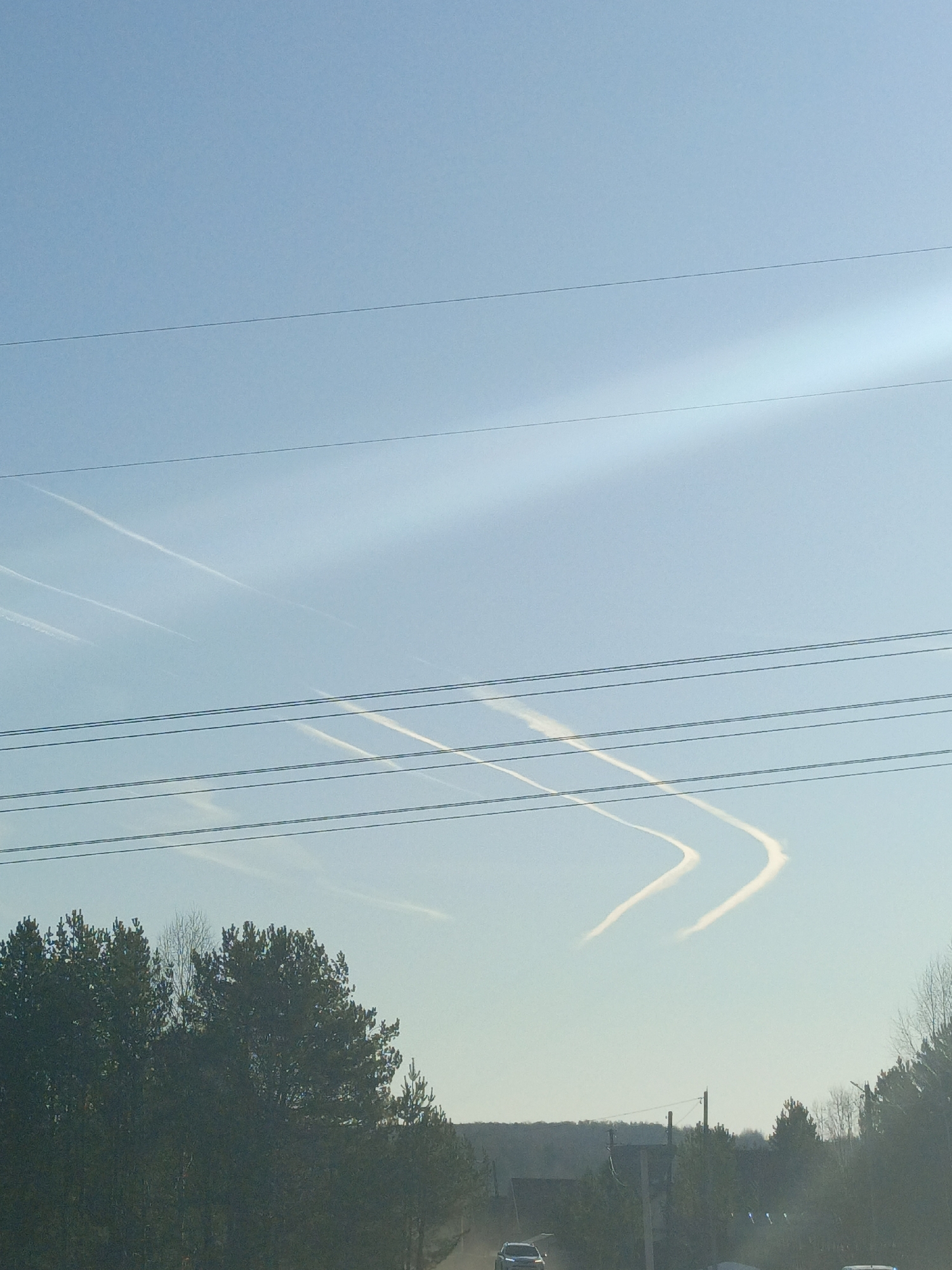 The image depicts a daytime view of the sky above Dubovaya, Russia. Contrails from airplanes are visible in the clear blue sky, some forming distinct V-shapes. Power lines stretch across the frame. Below, there's a line of dark green trees. In the foreground, cars are visible on a road. The overall scene suggests a rural or suburban environment.