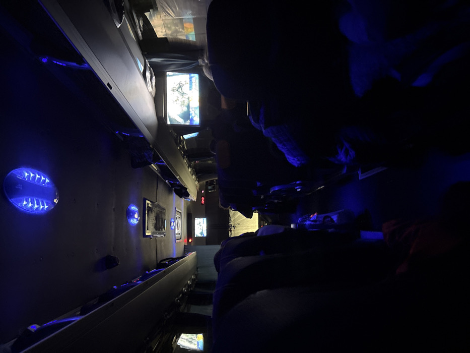The image depicts the interior of a passenger bus or coach under dim, predominantly blue ambient lighting. Numerous unidentifiable individuals are seated throughout the vehicle, appearing as dark silhouettes in the seats, facing forward.
**Environment:** The bus features a central aisle separating two rows of passenger seats. Overhead luggage compartments line both sides of the interior. The ceiling is dark, punctuated by several circular blue LED light fixtures that cast a strong blue glow onto the ceiling and parts of the aisle. Several small flat-screen monitors are mounted within the cabin; one prominent screen on the right displays what appears to be a broadcast or media content featuring a person's head and visible text. Another screen is visible on the left, and two smaller screens are further forward near the front of the bus. A red digital display, located above the central aisle towards the front, shows "22:12". Curtains or window coverings are drawn along the sides of the bus.
**People:** Passengers occupy many of the seats, their specific features obscured by the low light. They are stationary, presumably traveling. One passenger in the mid-ground on the right side appears to be wearing a light-colored or patterned garment on their upper body. A red object, possibly a bag or blanket, is visible on the floor near a passenger in the foreground on the left.
**Objects:** The seats are dark-colored. The blue ceiling lights have a distinctive internal finned or grid-like pattern. The floor of the aisle is dark, with subtle reflections of the blue light.
**Actions/Interactions:** The passengers are seated, but no specific actions or direct interactions are discernible due to the darkness and distance. Some individuals appear to be passively observing the screens.
**Location:** The image provides no visual information to confirm the specific geographical location within the city of Sedanovo, Russia. The scene is identifiable only as the interior of a bus.