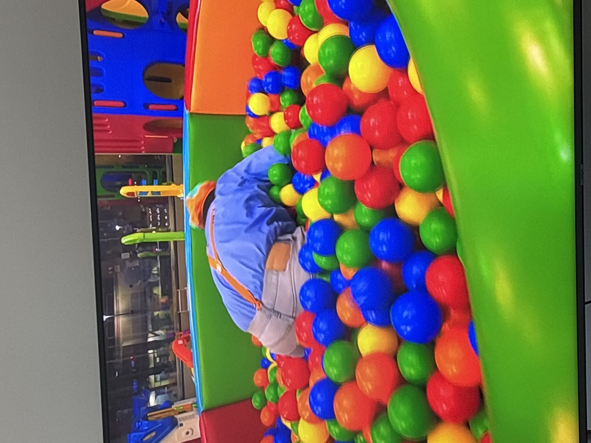 A person, dressed in a blue shirt and grey shorts with orange suspenders, is partially submerged in a ball pit filled with red, blue, green, and yellow balls. The ball pit is part of a colorful indoor playground with red, blue, and yellow plastic structures visible in the background. The scene appears to be indoors, possibly in a children's entertainment center.