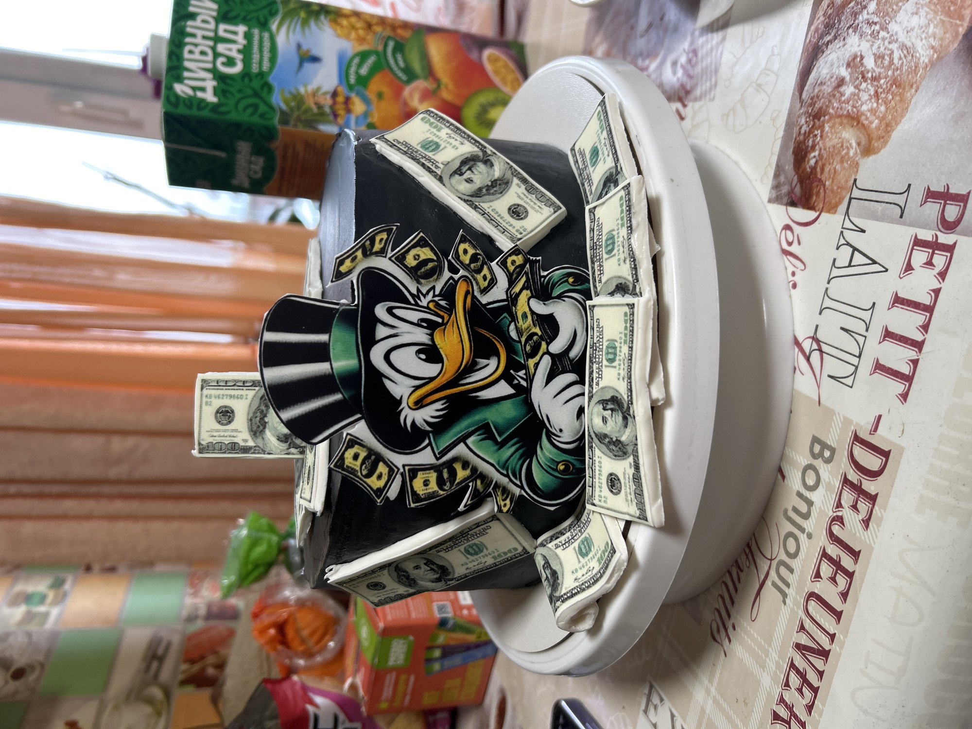 The image shows a single-tier, round cake with black frosting, positioned centrally on a white cake stand. The cake is decorated with edible elements, including a cartoon character resembling Scrooge McDuck, dressed in a green suit and black top hat, depicted holding and surrounded by falling $100 bills. Edible $100 bills are also affixed to the sides of the cake, with additional bills arranged flat on the white cake stand around the cake's base. One folded $100 bill stands upright on top of the cake. The cake rests on a light-colored tablecloth patterned with plaid and French text such as "DEJEUNER" and "Bonjour," along with a printed image of a pastry. In the background, out of focus, a patterned wall is visible on the left, alongside a pink cup, various packaged items, and a partial view of a smartphone. On the right, a large window draped with sheer orange curtains is visible, next to a green carton of "ДИВНЫЙ САД" brand tropical fruit juice. The scene is an indoor domestic setting in the city of Ольховка, Russia. No people are present.