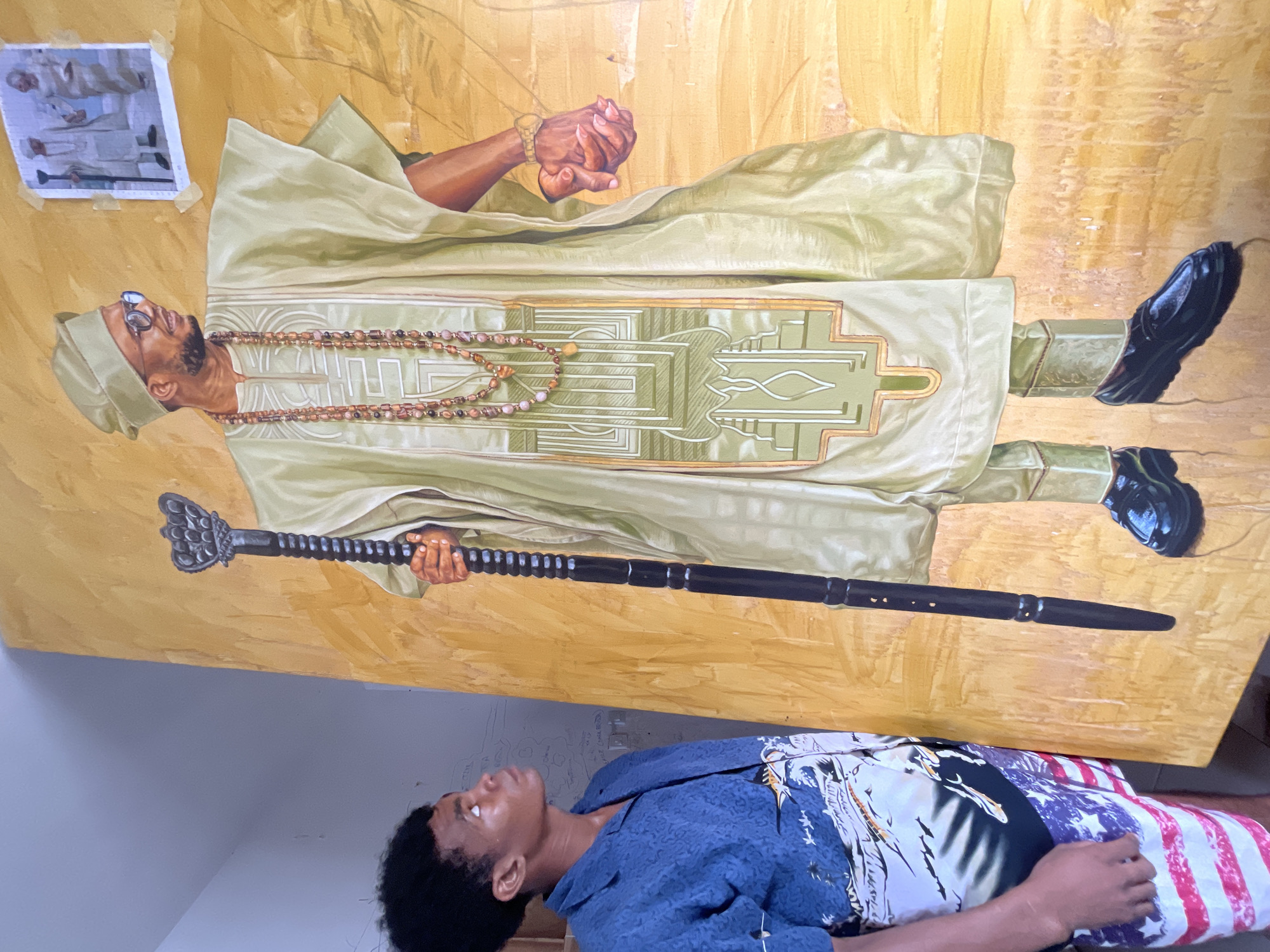 A young man with dark, curly hair stands to the left of a large, vibrantly painted portrait of a man in traditional Nigerian attire. The portrait depicts a man wearing a light green agbada and a matching fila, adorned with intricate beading and a gold watch. He holds a dark, ornate staff in his left hand and clasps his right hand in front of him. A small framed photograph of two figures in similar traditional dress is taped to the background of the portrait, near the upper right corner. The young man is wearing a blue patterned short-sleeved shirt and shorts with an American flag motif. The background of the scene appears to be an indoor space, possibly a studio or workshop, with a light-colored wall on the left and a wooden surface visible behind the young man. The overall lighting suggests a bright, natural light source from the right. The location is identified as Ado, Nigeria.