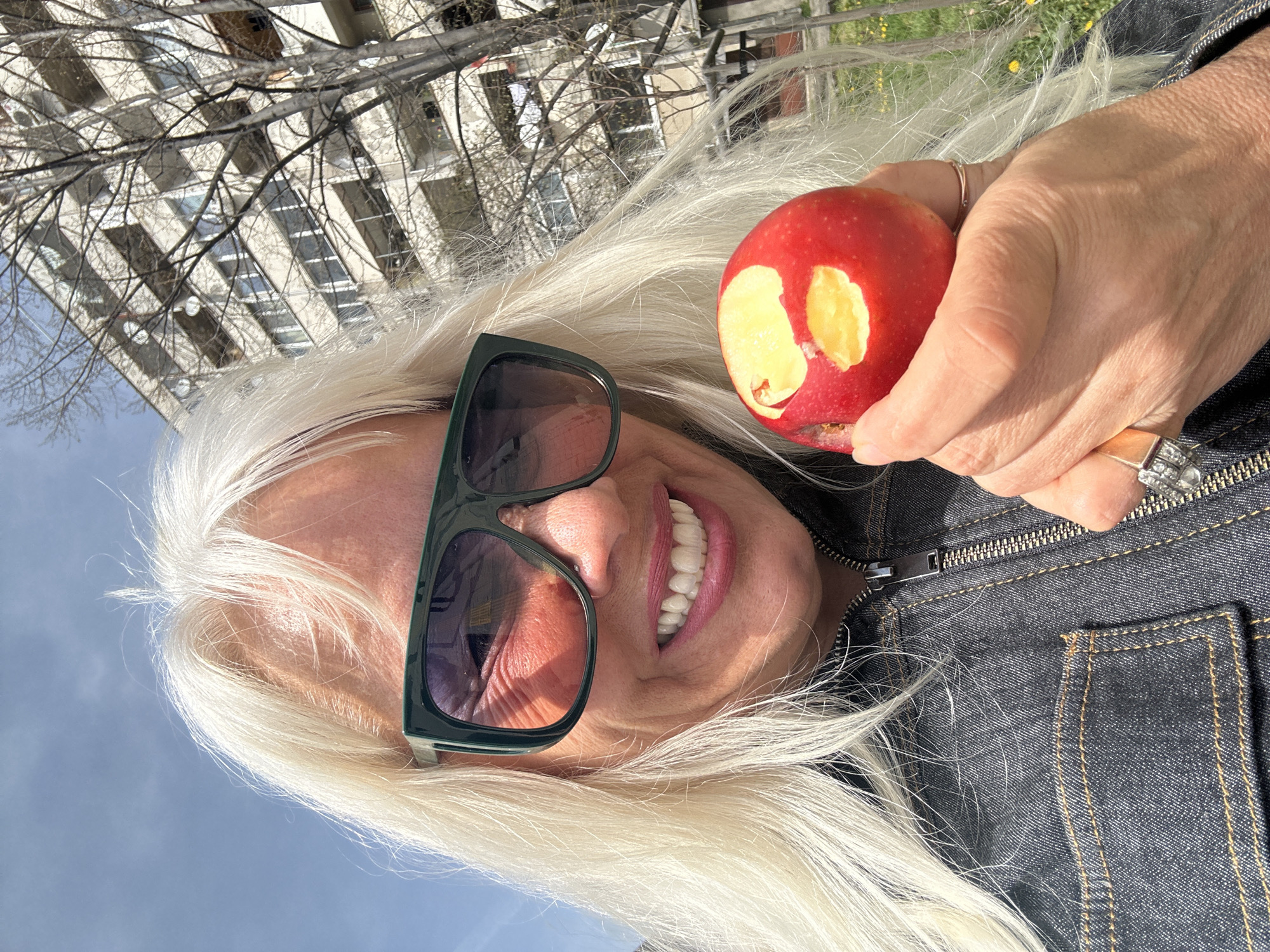 A woman with long, white hair, wearing dark green sunglasses and a dark denim jacket, is holding a partially eaten red apple in her right hand. The apple has two visible bite marks. She is smiling, showing her teeth. She is wearing a ring on her right hand. In the background is a building with multiple windows and bare tree branches against a pale blue sky. This location might be in the city of Sofia, Bulgaria.