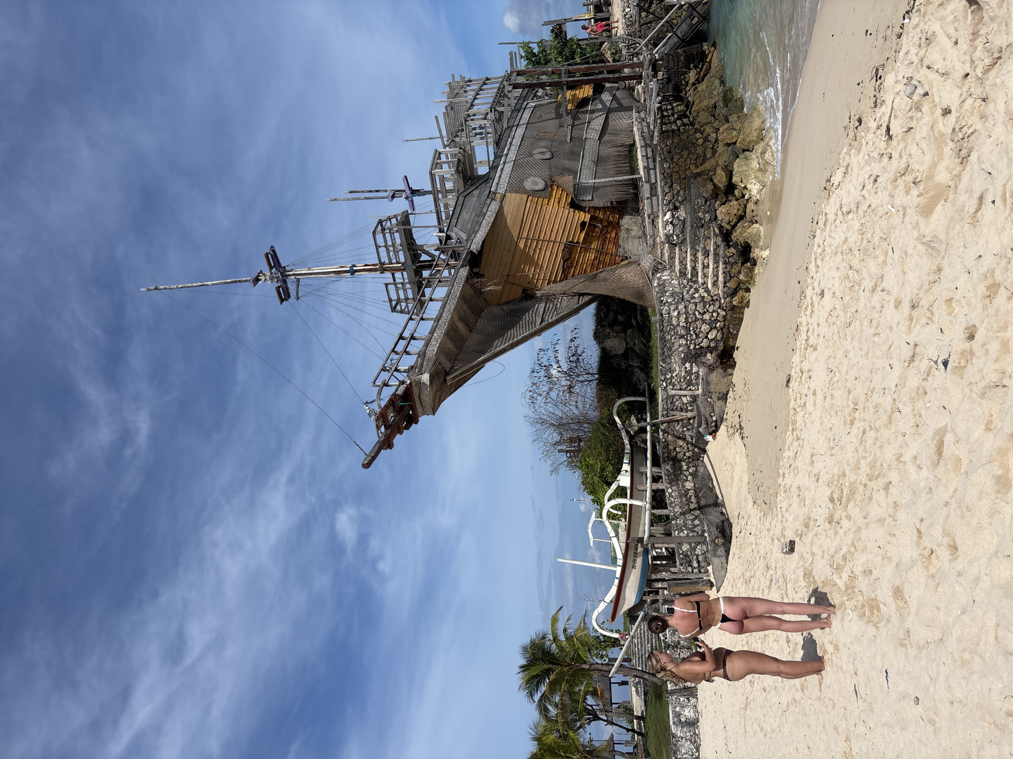 The image shows a partially constructed wooden ship, possibly a replica, situated on a rocky outcrop adjacent to a beach. The beach consists of light-colored sand. Two women stand on the beach, viewed from the side. Palm trees are visible in the lower left corner of the frame. A small boat sits between the women and the ship. The sky is blue with scattered clouds. Based on the presence of the ship replica, this scene is likely in the city of Kuta Selatan, Indonesia.