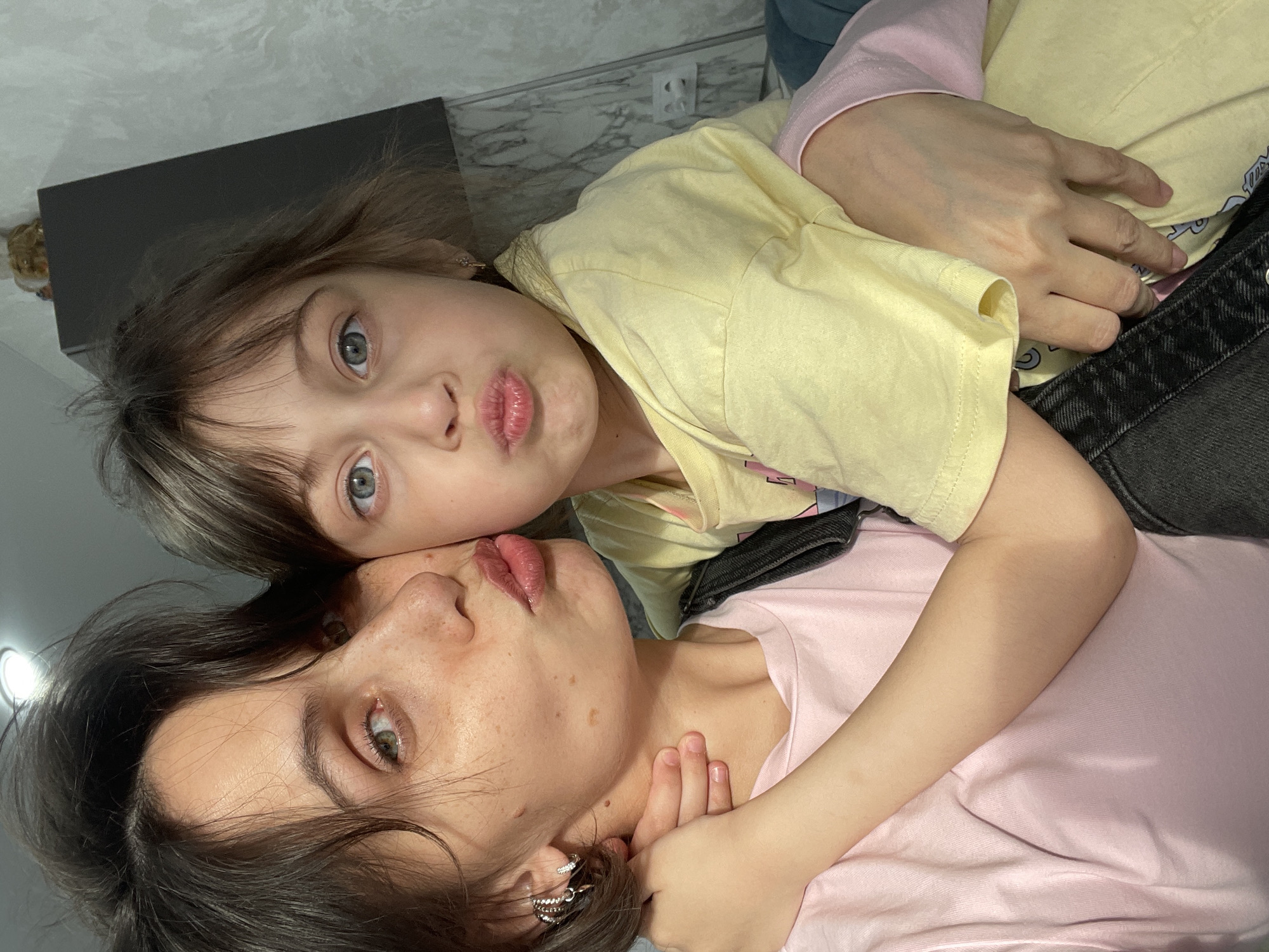 Two people, likely a young child and an adult female, are lying down and posing for a photo. The child, wearing a yellow shirt, is on top, with her face close to the camera and lips puckered. She has light hair and blue eyes. The adult female is below her, wearing a pink shirt. Her face is also close to the camera, with lips puckered as well. She has dark hair and freckles. The environment appears to be indoors, with a gray wall and an electrical outlet visible in the background. There is also a dark rectangular object in the upper left corner, possibly a piece of furniture. I have no evidence to suggest this photo was taken in Izhevsk, Russia.