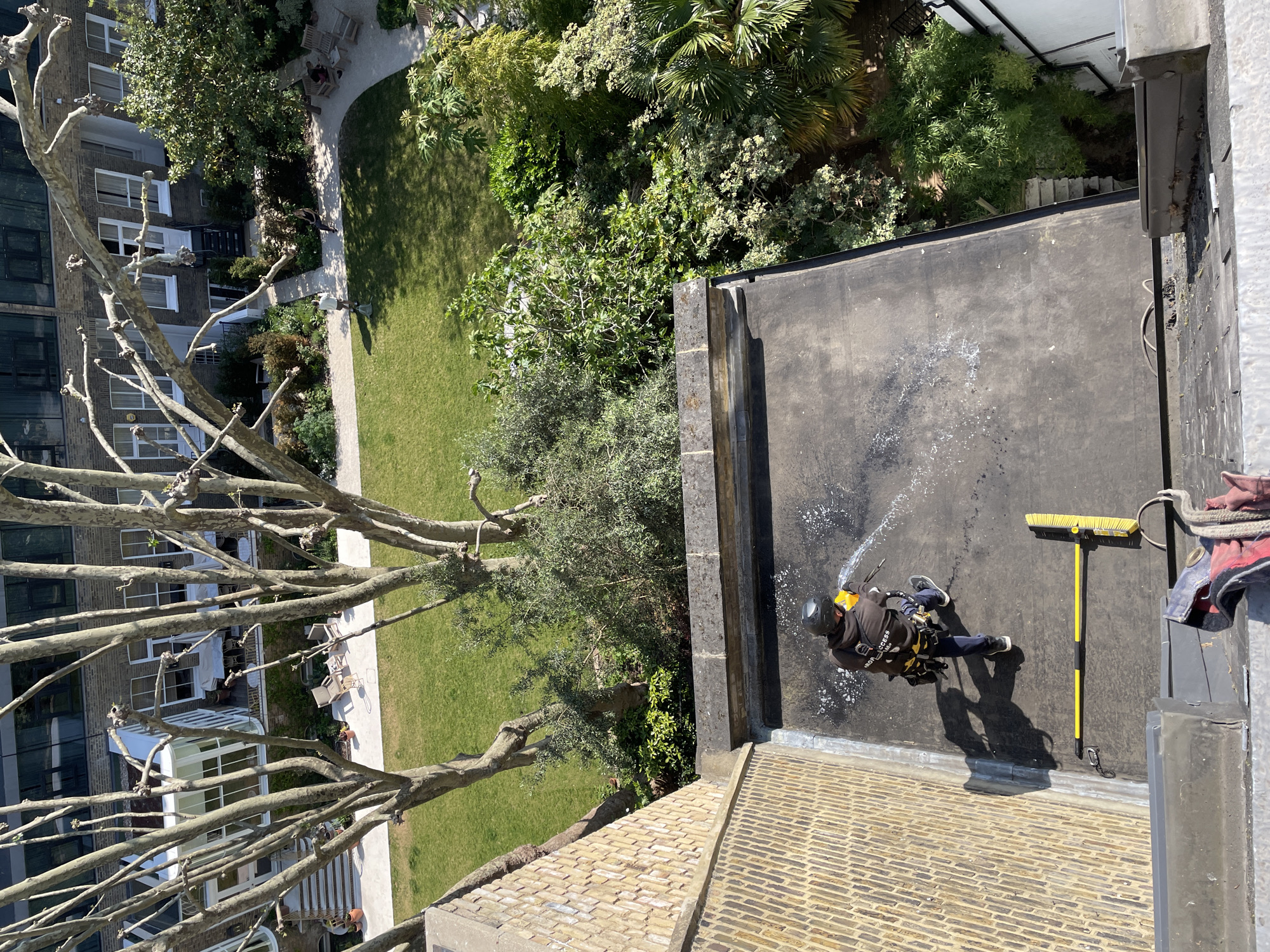 An aerial view shows a man cleaning a flat rooftop in London, United Kingdom. The man is wearing a dark-colored jumpsuit, helmet, and safety harness, using a water hose and squeegee on the black roof. Adjacent to the roof is a residential building of brown brick and an open space of lawn and garden. The environment is bright and sunny.