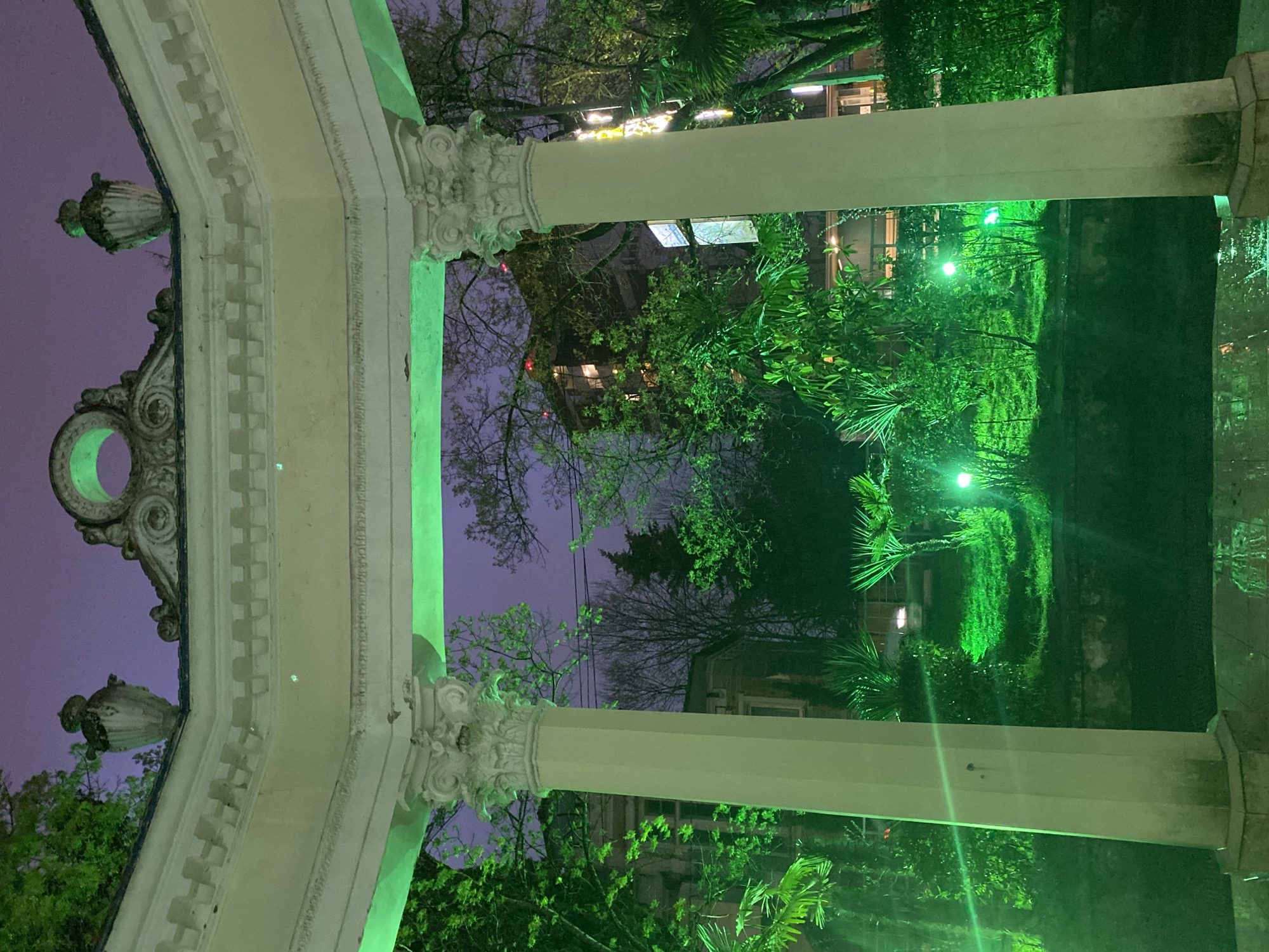 The image depicts a green-lit arbor at night, likely in Sochi, Russia. The arbor's architectural elements include white columns and ornate details such as urns. Through the arbor's opening, various trees and buildings are visible. The scene is illuminated by green lighting, which gives the foliage and the arbor an eerie glow.