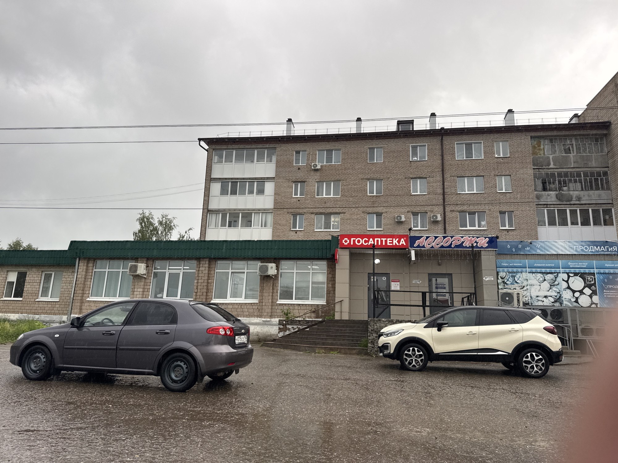 This media file captures an outdoor urban scene featuring two cars parked on a wet asphalt surface in front of a multi-story brick building under an overcast sky. The overall impression is one of a cloudy, possibly rainy day.
The primary subject is a four-story brick apartment building with numerous windows, some of which feature white frames and external air conditioning units. The upper part of the building has multiple balconies, some enclosed, others open and appearing somewhat dilapidated. Below the main building, a single-story structure with a dark green sloped roof extends outwards. This lower building also features large windows and air conditioning units.
Two cars are prominently visible: a dark gray sedan (appears to be a Chevrolet Lacetti) parked on the left, facing away from the viewer, and a cream-colored SUV with a black roof (likely a Renault Kaptur) parked on the right, facing left. Both vehicles are on a visibly wet ground, suggesting recent rain. Utility lines are stretched across the upper portion of the frame.
Several signs are affixed to the buildings. A prominent red sign reads "ГОСАПТЕКА" (GOSAPTEKA), indicating a state pharmacy. Next to it, a blue sign displays "АССОРТИ" (ASSORTI), likely a shop name. Further right, another blue sign reads "ПРОДМАГИЯ" (PRODMAGIYA), which translates to "Food Magic," indicating a grocery store. Below "ПРОДМАГИЯ," smaller text is visible: "ТОРГОВАЯ СЕТЬ" (TORGOVAYA SET' - Trading Network), "Товары, доступные каждому" (Goods available to everyone), "Домашняя выпечка" (Homemade pastries), and "Продукты местных производителей" (Products from local manufacturers).
No people are visible in the image, and there is no discernible activity taking place. The time of day appears to be daytime, given the ambient light, though obscured by cloud cover. The Russian text on the signs confirms the location context of Mozhga, Russia.