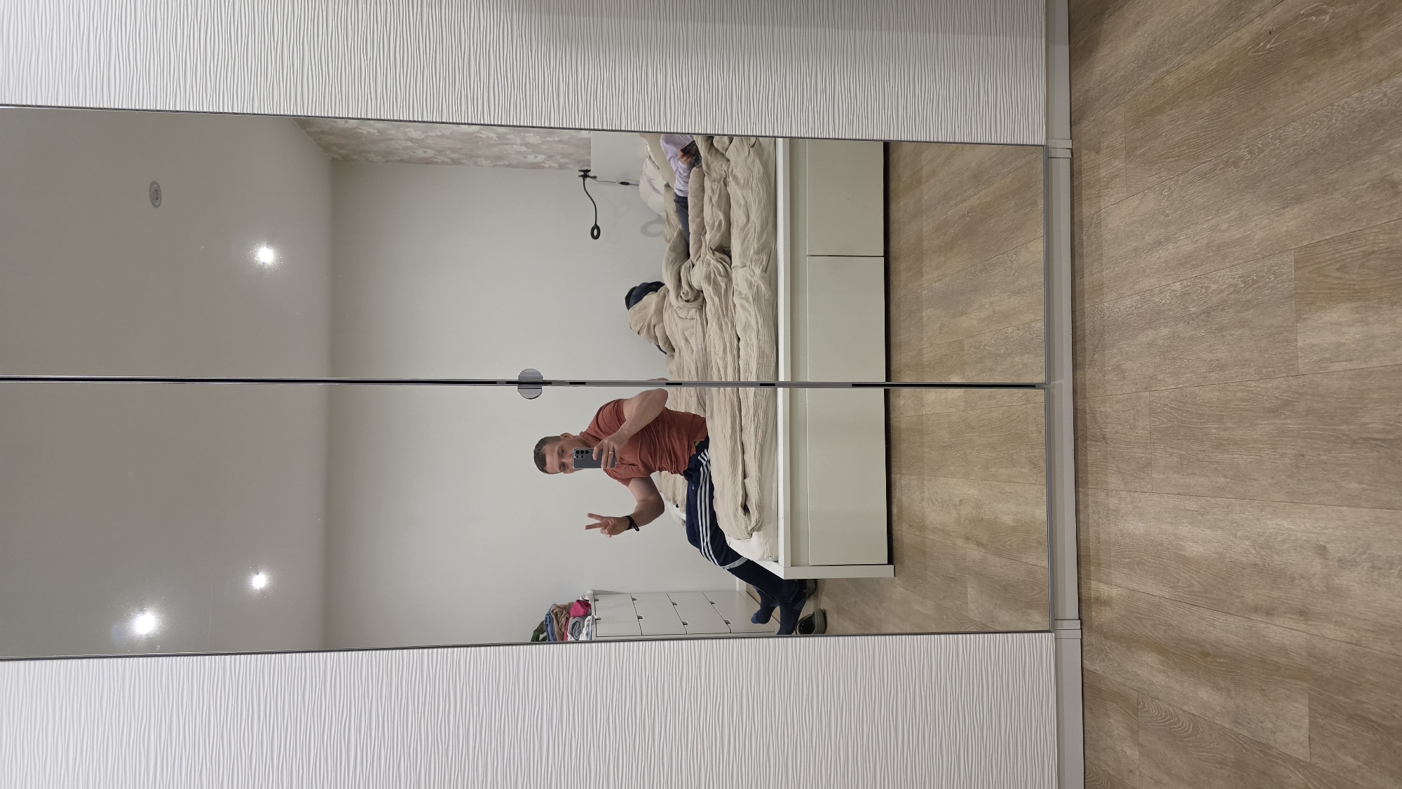 The image shows a reflection in a mirrored closet. A man is taking a selfie in the mirror. He is wearing a reddish-brown t-shirt and black track pants with white stripes. He is making a peace sign with his left hand. The reflection also shows part of a bed with a beige comforter, a white dresser with clothes on top, and a light-colored wooden floor. The top and bottom of the closet doors have a textured white panel. The location is a bedroom in Ihringshausen, Germany.