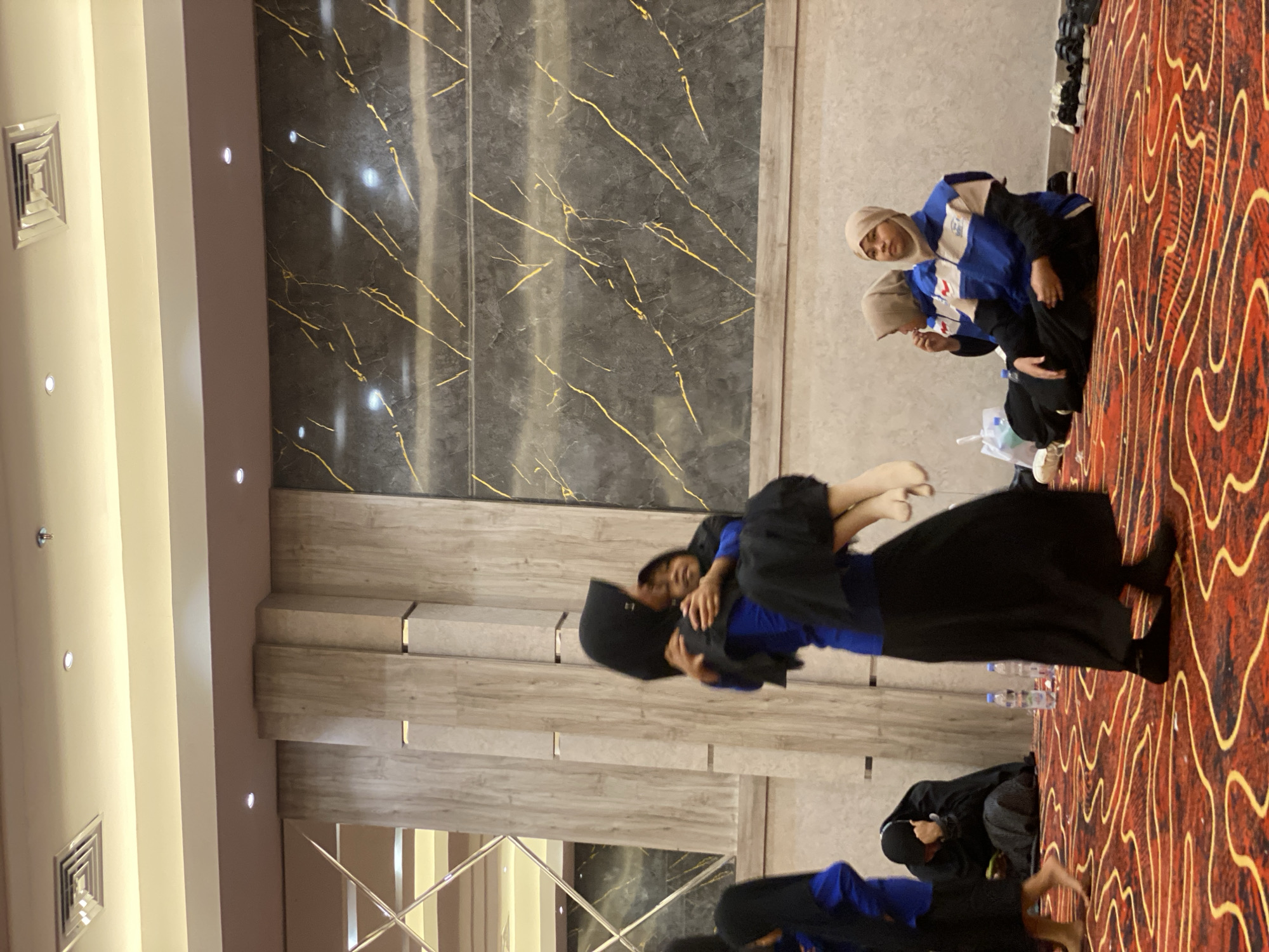 The image depicts an indoor gathering at the Borneo Convention Centre (BCC) in Banjarbaru, Indonesia. In the foreground, a person wearing a blue top and black skirt is carrying a child, also in a black hijab, in their arms. The child's legs are visible, and they appear to be an infant or a very young child. To the left, another person in a blue top and black skirt is partially visible. In the mid-ground, two individuals are seated on the floor. One, wearing a black hijab, is hunched over with their hand near their face. The other, also wearing a hijab, is in a blue and beige jacket and black pants, looking towards the right. Several water bottles and a white plastic bag are on the floor. The floor is covered with a red carpet featuring a gold wavy pattern. The background features a wall with vertical wooden panels and a section with dark grey marble and gold veining, typical of the BCC's interior design. The ceiling has a metallic ventilation grate and recessed lighting. The overall scene suggests a cultural event, performance, or religious gathering.