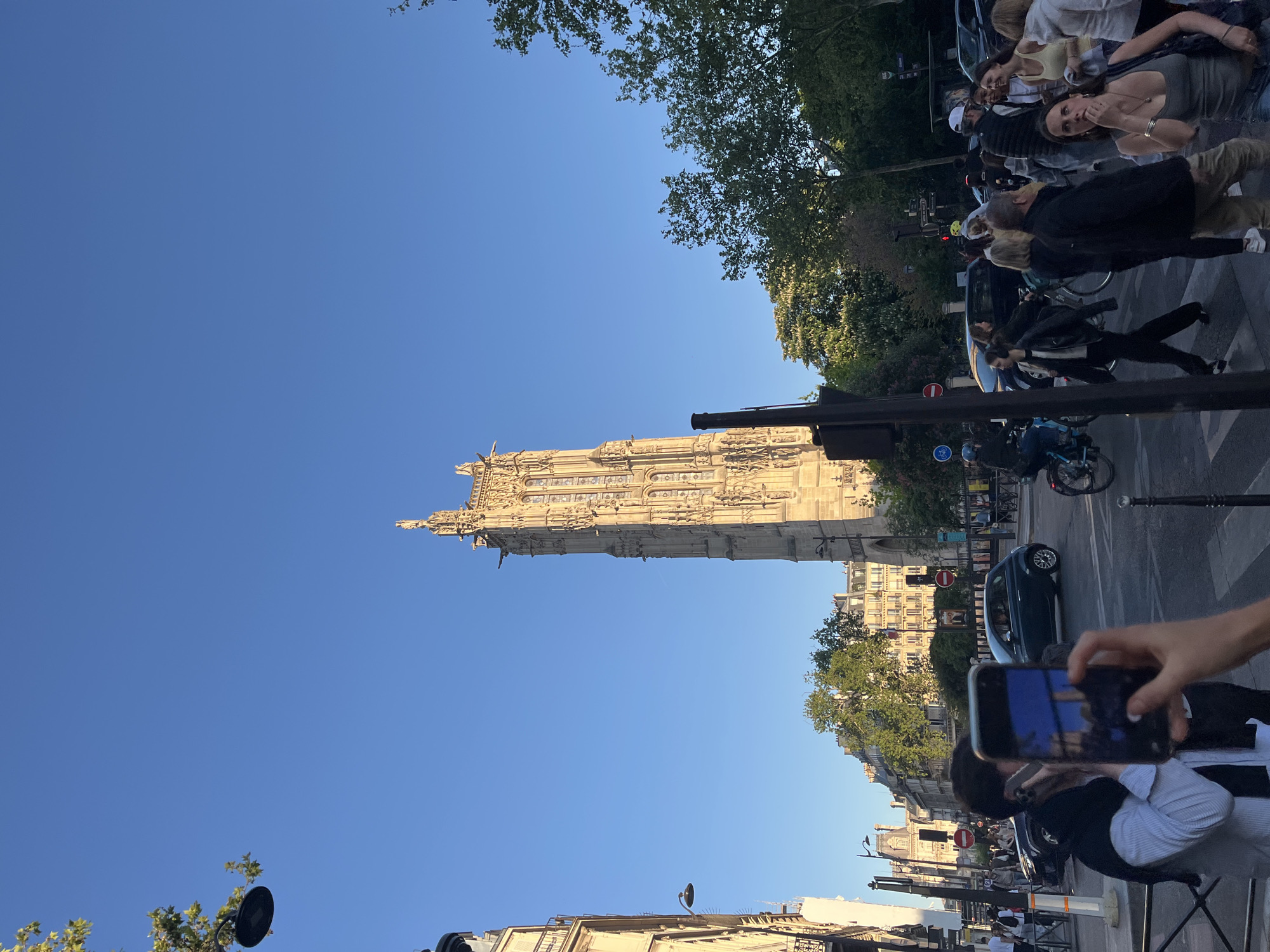 The image captures the Tour Saint-Jacques in Paris, France, viewed from street level. The tower, a notable example of Flamboyant Gothic architecture, rises against a clear blue sky. The lower part of the image shows Parisian street elements, including buildings, street signs, vehicles, and pedestrians. A person is holding a smartphone, presumably taking a picture of the tower. Several other people are standing on the sidewalk.