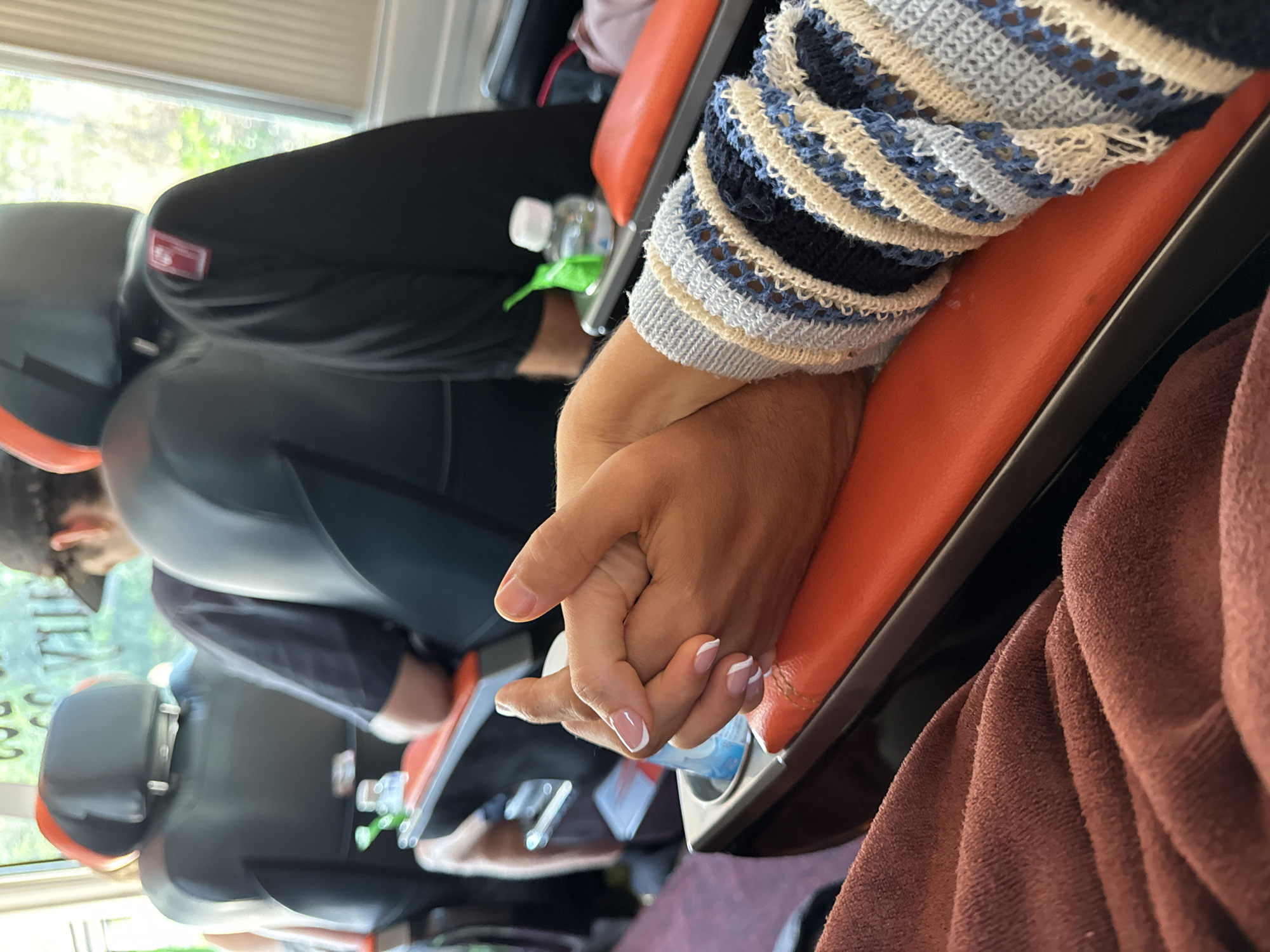 The image captures an intimate moment inside a passenger vehicle, likely a bus or van, during daytime travel in Phan Son, Vietnam. In the foreground, two hands are clasped together, symbolizing affection or comfort between two individuals. One arm, partially visible, has a darker skin tone and is covered by a patterned knit sleeve featuring blue, white, cream, and black stripes. The other hand, with a lighter skin tone and neatly manicured nails with white French tips, is held gently.
The setting is an interior space dominated by orange and black seats, typical of passenger transport. Several other passengers are visible in the midground and background, sitting in similar seats, though their faces are largely obscured or blurred. One person in the background wears a black cap. Small plastic water bottles are visible tucked between seats. A brown or maroon textured fabric, possibly a blanket or clothing, occupies the far right of the frame.
Bright natural light streams in from a large window on the left side, indicating daytime. Outside the window, a blurred view of lush green foliage and a bright sky suggests an outdoor, possibly rural or scenic, environment. Window blinds are visible at the top of the window. A small, red rectangular label with unidentifiable white text or a logo is attached to the back of a seat headrest. The overall scene depicts travel and companionship.
