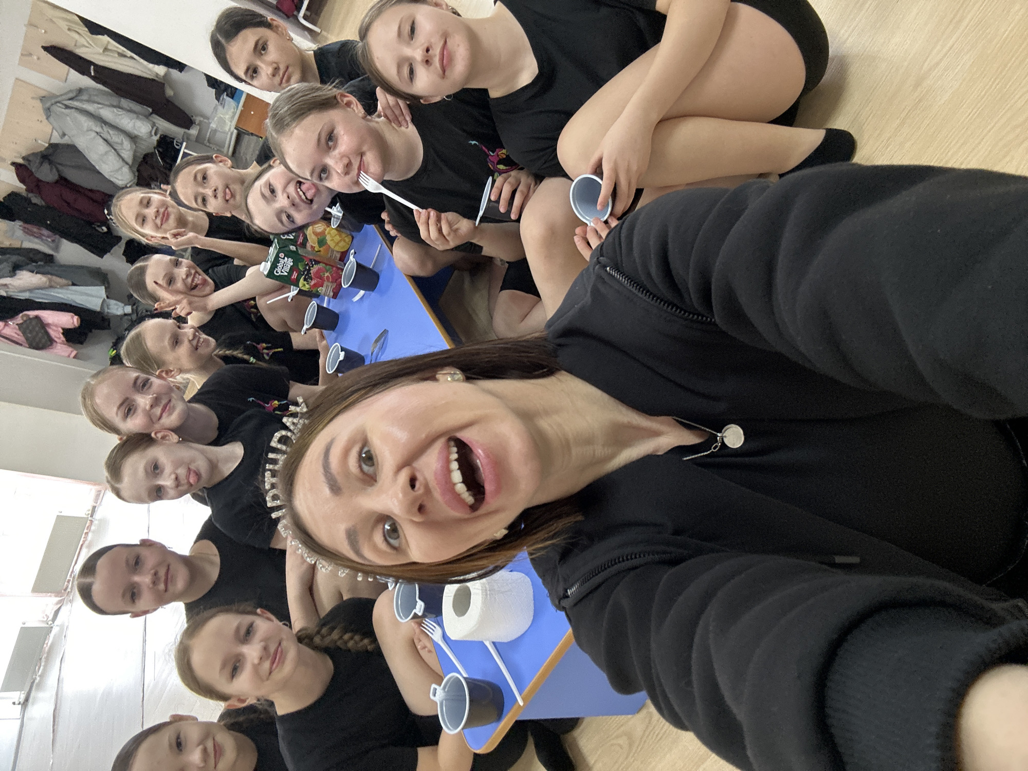 A group of about twelve young girls and an adult woman are posing for a selfie in what appears to be a classroom or studio. All are Caucasian. The girls are sitting around a small blue table with blue cups and white forks set on it. One juice box is visible on the table. The woman is in the foreground wearing a black hoodie and a "birthday" tiara. The girls are dressed in black t-shirts and some have black shorts. The background shows a wall with a clothes rack and some clothes hanging on it. The floor appears to be made of wood. There is no definitive information in the photo to confirm the location is Votkinsk, Russia.