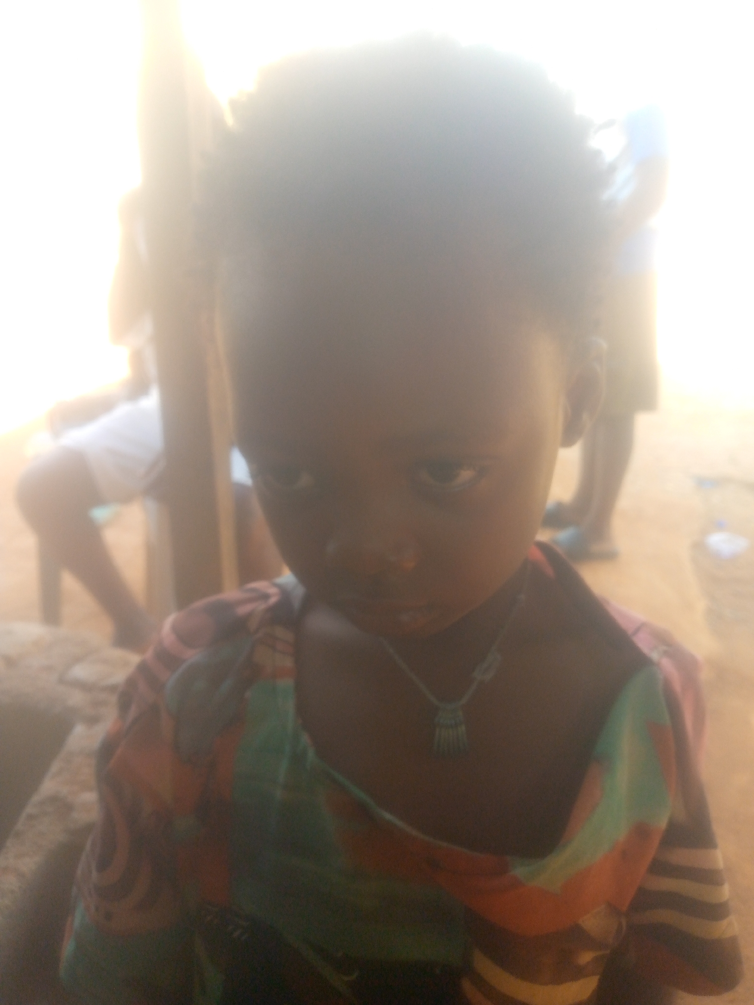 A close-up photograph depicts a young girl with dark hair, wearing a colorful patterned shirt and a necklace with a small pendant. The photo is taken in an outdoor setting in the city of Unknown, Unknown. There are partially visible figures of people in the background, along with some chairs. The lighting appears to be bright, potentially due to sunlight.