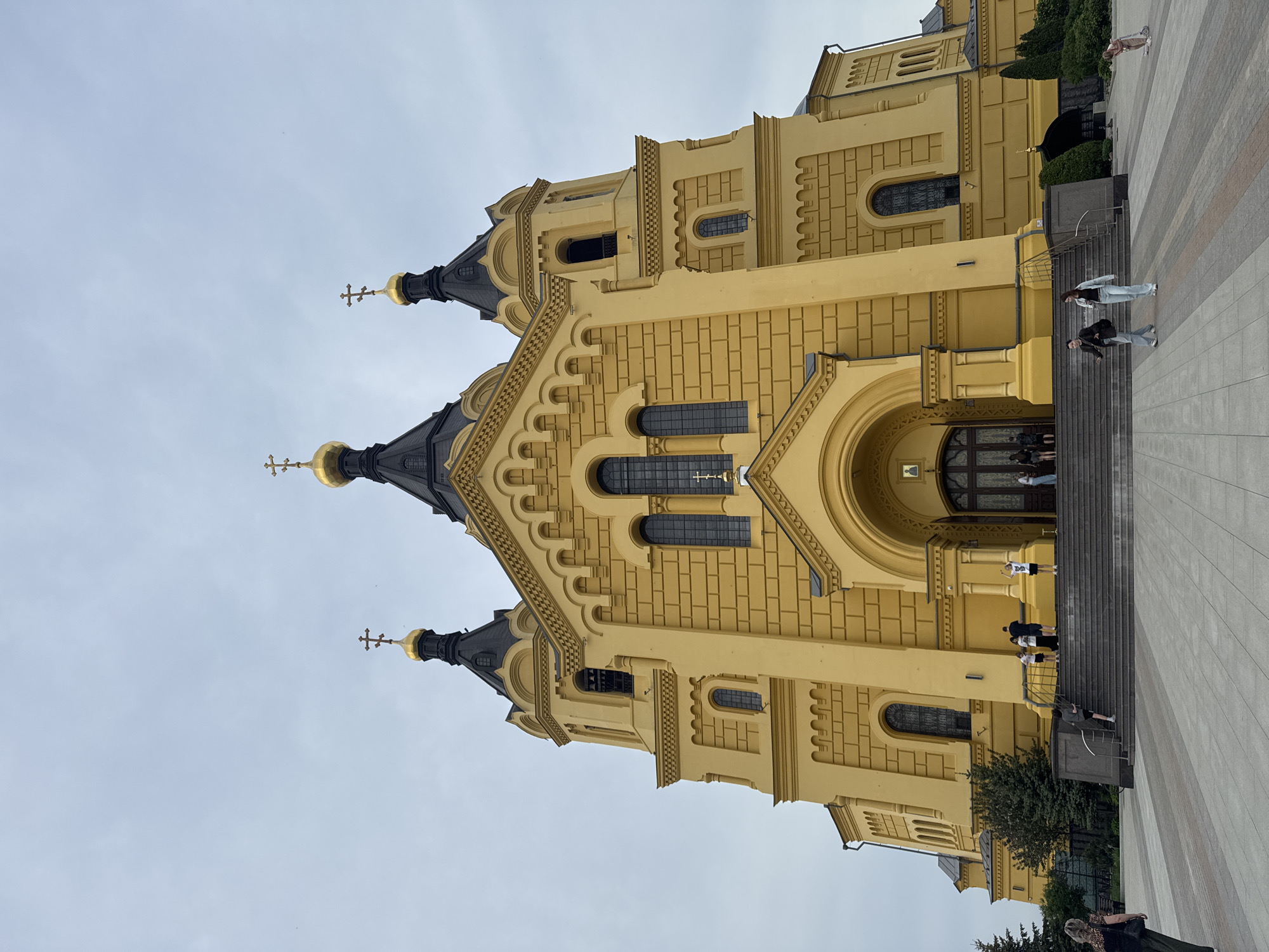 The image shows the Alexander Nevsky Cathedral in Nizhny Novgorod, Russia. The cathedral is a large, yellow building with several black domes topped with golden crosses. The architecture has a brick-like pattern on the exterior walls. A set of stairs leads to the entrance of the cathedral. Several people are on the stairs and in the surrounding area. The sky is overcast. The ground is paved with concrete. Trees and other greenery are visible near the base of the building.
