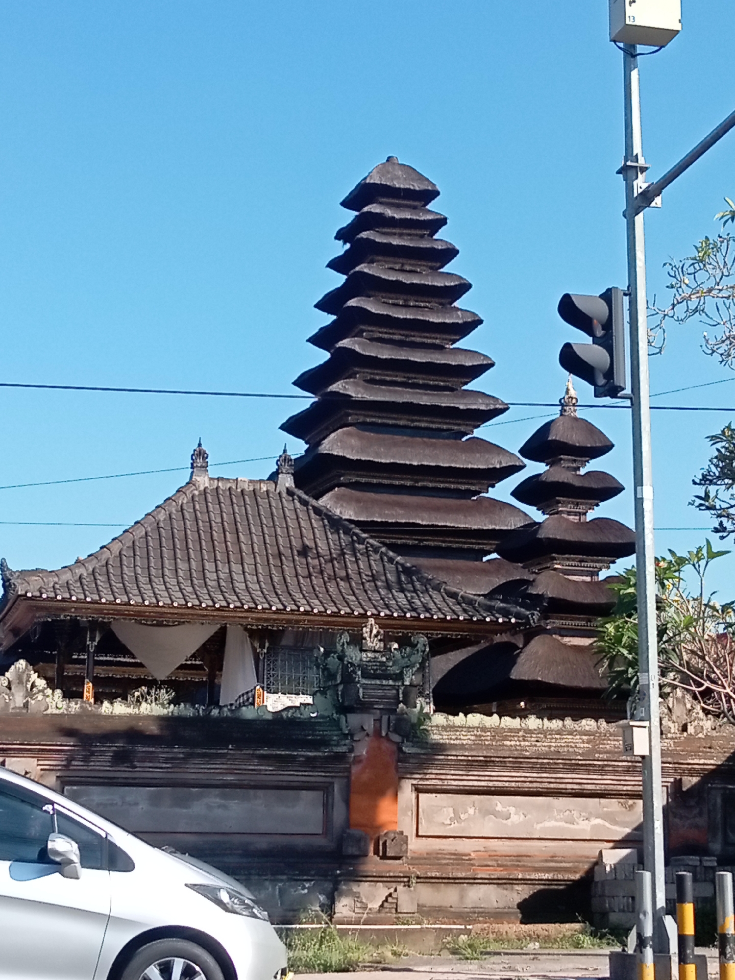 A low-angle, outdoor, daylight shot captures a multi-tiered, dark thatched-roofed structure resembling a pagoda or temple against a clear blue sky. The structure is elaborate, with many layered roofs that decrease in size as they ascend. In front of this structure is a dark, weathered stone wall with an orange-tinted section on the left. To the right of the main structure, a traffic light is visible on a pole, with a small white box attached to the top of the pole. The base of the pole has yellow and black stripes. In the foreground, the front of a silver car is partially visible, parked on the left side of the frame. The environment appears to be urban, with overhead wires and sparse vegetation visible to the right of the traffic light. The overall scene suggests an architectural landmark in an open area, possibly a public space or street.