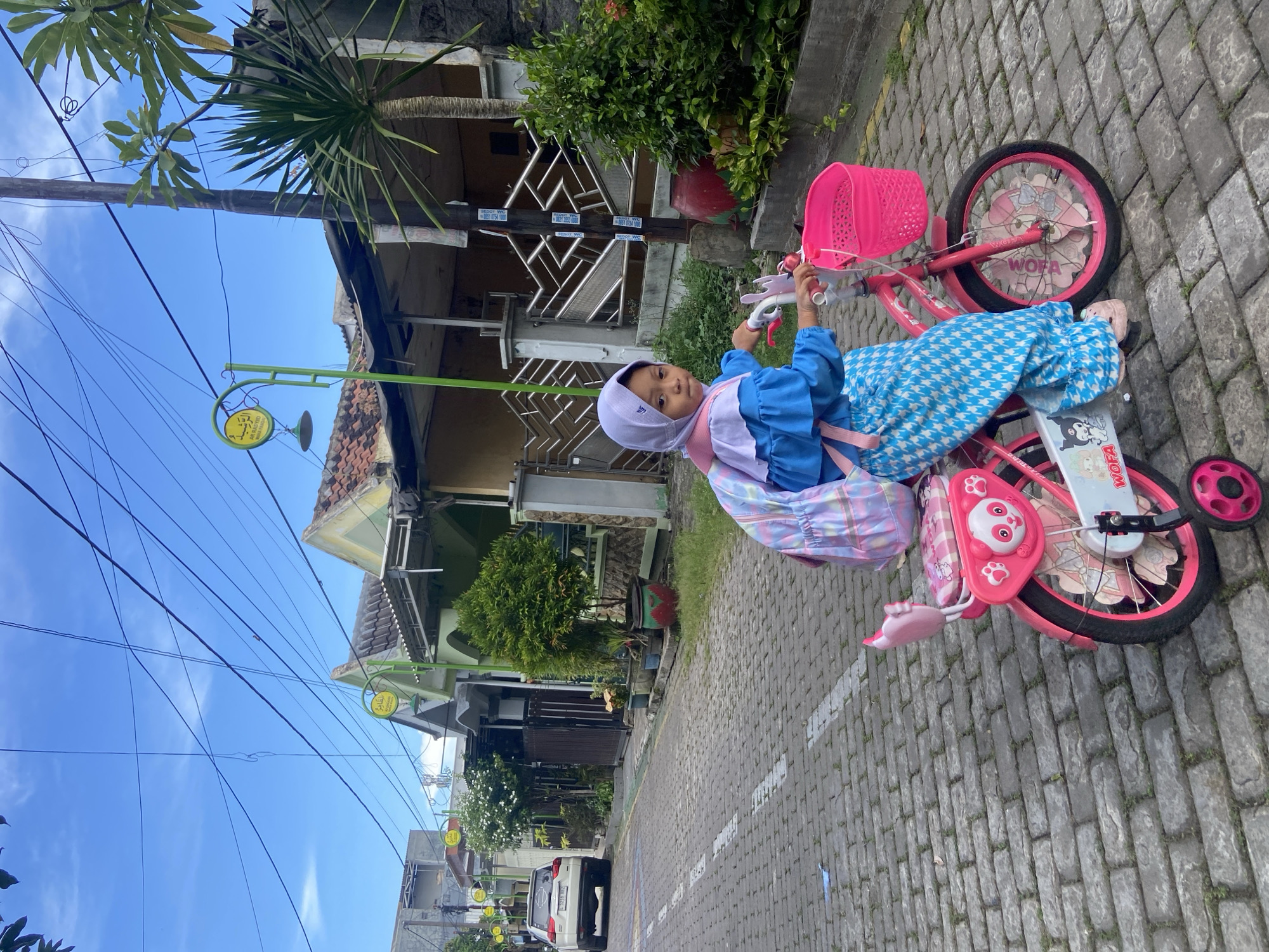 A young girl is seen sitting on a pink bicycle with training wheels on a paved residential street in Gelam, Indonesia. She is wearing a light purple hijab, a blue long-sleeved top, and blue and white patterned pants, along with a pastel-colored backpack. She looks towards the camera with a calm expression, holding the handlebars.
The bicycle is brightly colored in pink, featuring a front basket, a wheel cover with a bow design and the text "WOFA," and a sticker that appears to read "Kuroda." Panda-themed decorations are also visible near the pedals and front wheel.
The setting is a residential area with houses on both sides of the cobblestone street. Green plants and trees line the street and surround the houses. The sky is bright blue with a few clouds, indicating a sunny day. Numerous black power lines crisscross overhead. Decorative street lamps with circular yellow signs framed in green are visible. One such sign clearly displays the text "AGEN PULSA & KUOTA" (Pulse & Quota Agent), "WA: 0857 5854 5060," and some Arabic script. A white car is parked further down the street in the background.