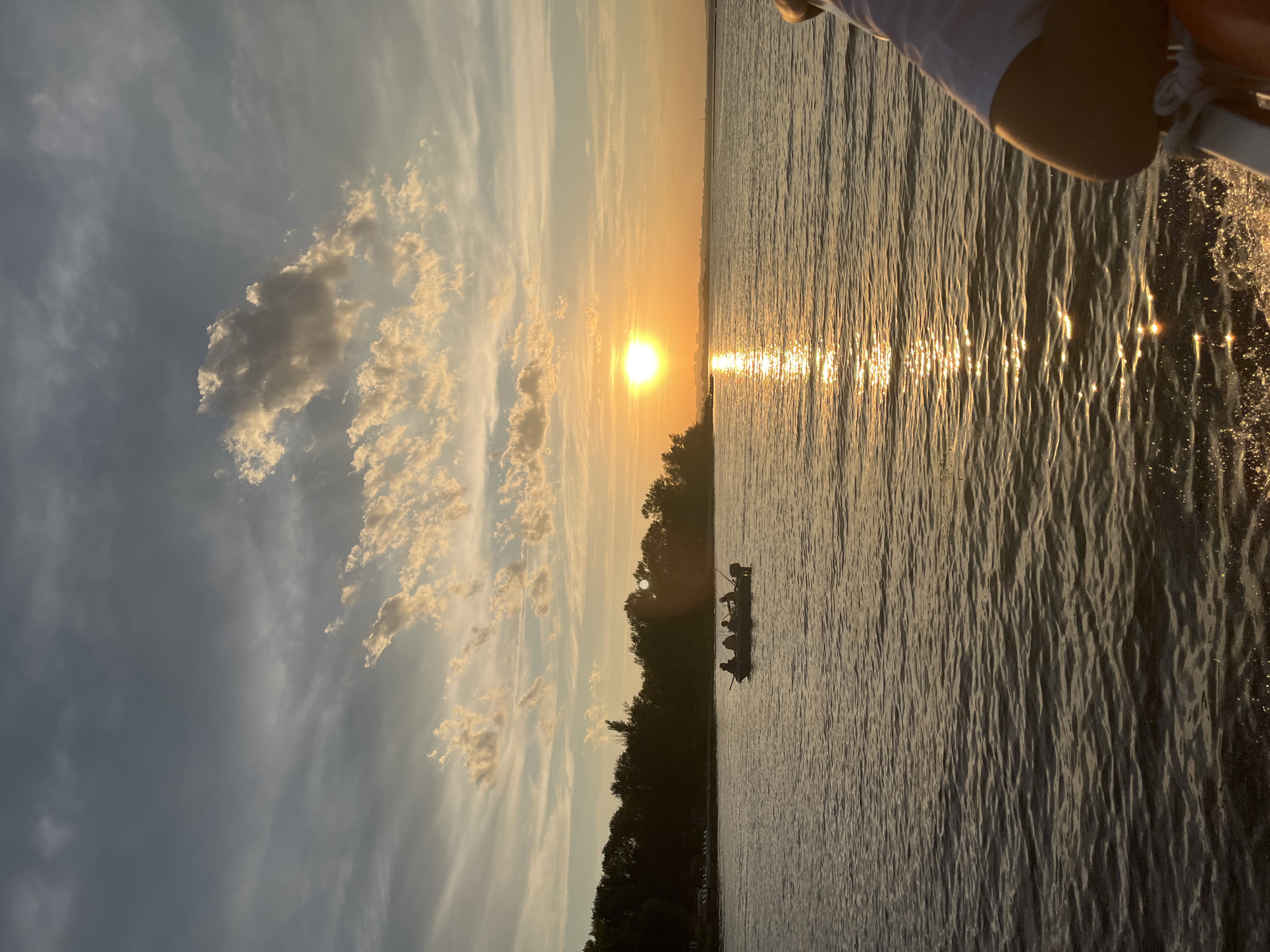 In Shumeika, Russia, a group of five individuals are seated in a small motorboat on a calm body of water during sunset. The sky above is a canvas of dusky blues and grays, interspersed with scattered, wispy clouds catching the golden light of the setting sun. The sun, a bright orb, casts a shimmering reflection across the water, creating a path of light that stretches towards the viewer. A dark, tree-lined shore forms the background of the scene. The overall impression is one of peaceful passage on the water as day transitions into evening.