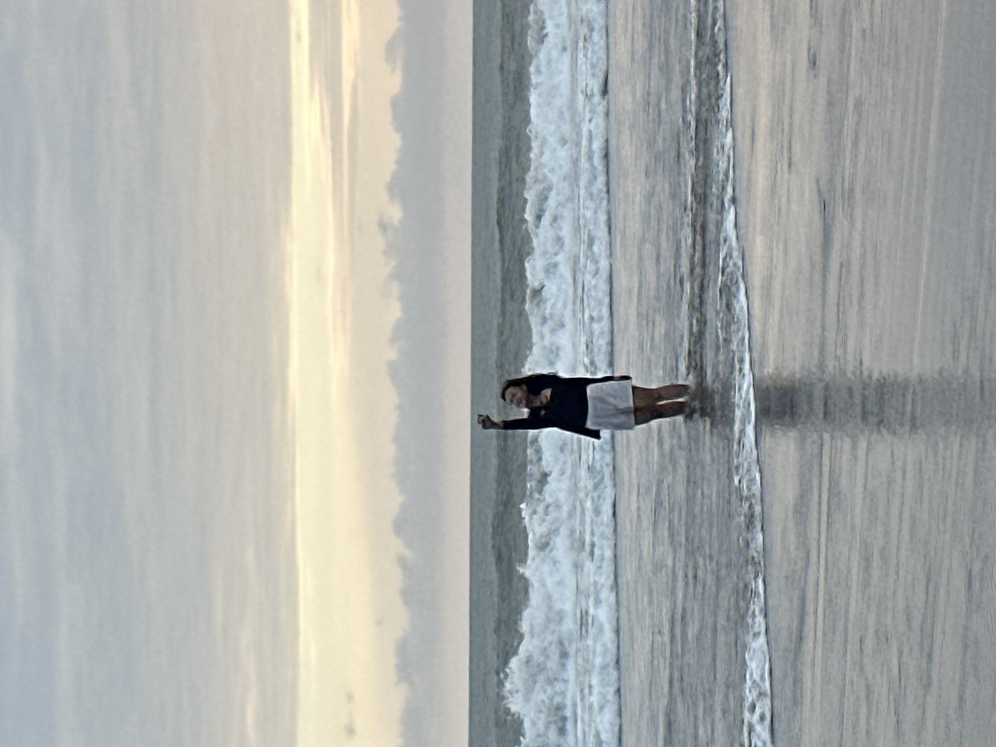 A person is standing in the ocean on a beach in North Kuta, Indonesia. The person is wearing a black jacket and white shorts. They are holding up their right hand, and it appears they are taking a selfie. The ocean has waves, and the sky is cloudy with a hint of yellow and orange from the sunset. The overall atmosphere is serene and peaceful.