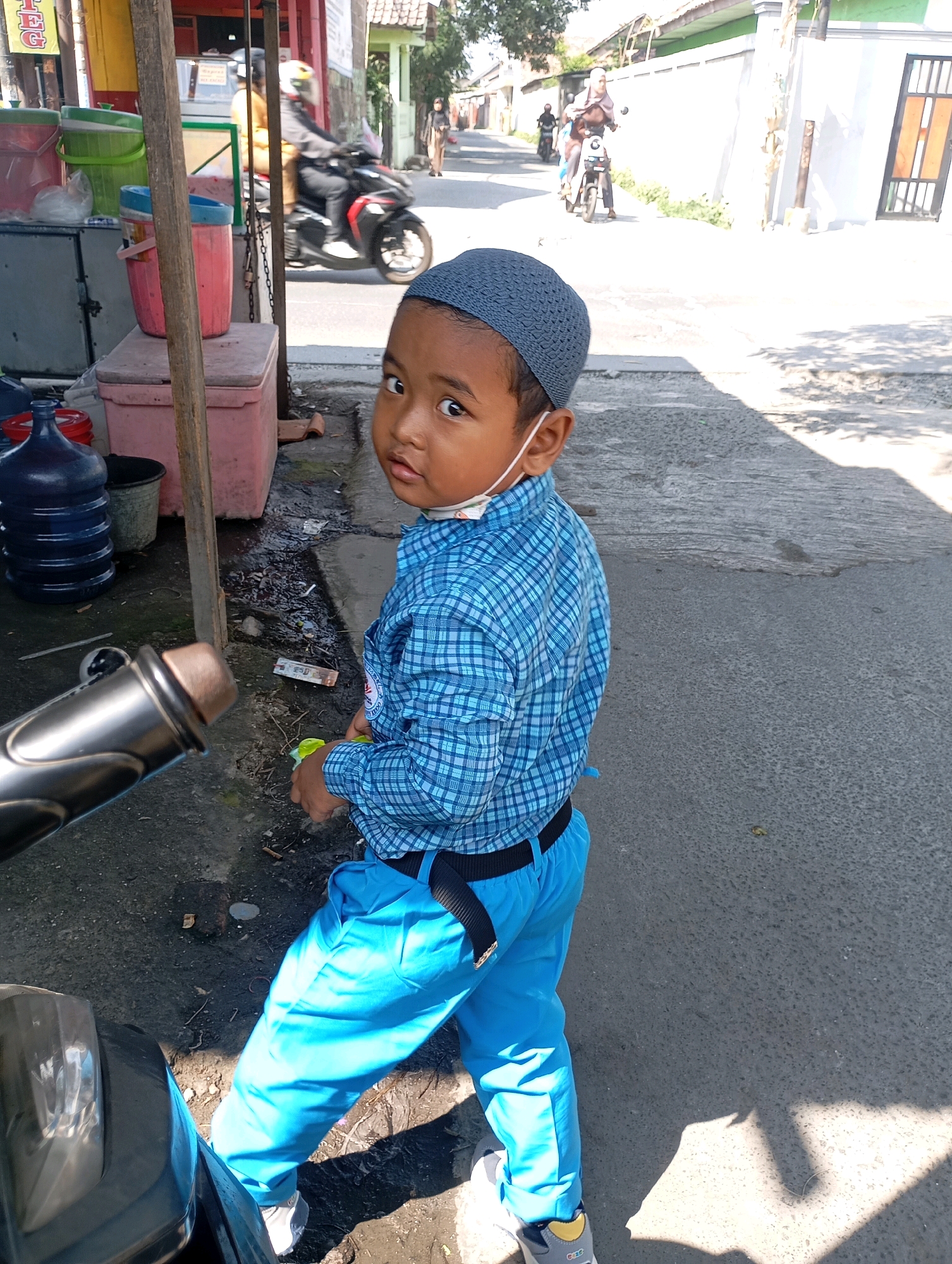 In Babelan, Indonesia, a young boy wearing a blue checkered shirt, blue pants, and a blue skullcap stands on a paved street. He is partially obscured by the front of a dark-colored motorcycle and is looking to his left. A white mask is visible on his face. To his left, a wooden stall with various containers, including large blue water jugs, is set up. In the background, several people are visible on motorcycles and walking along the street, with buildings and trees lining the area. The sun is casting shadows on the ground.