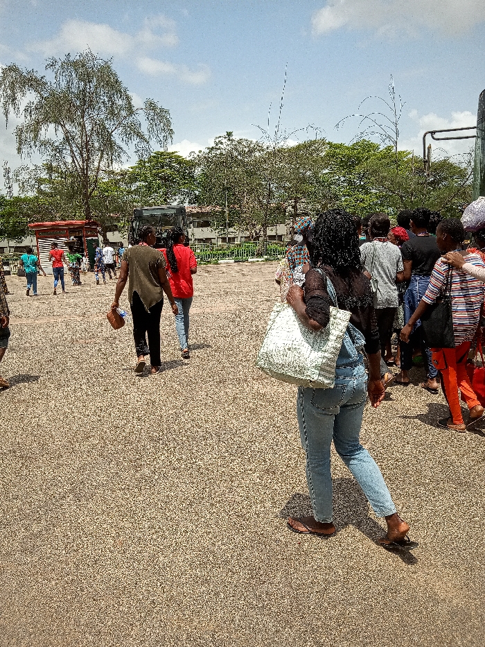 The image shows a group of people in a paved area. They are standing and walking near a bus. One person is wearing a red shirt and walking toward the left side of the image. Another person is carrying a large white bag and walking toward the right side of the image. The image is taken from a slightly elevated perspective. The location of the image, as determined by the coordinates, is Ota, Nigeria. There are trees on the periphery and a single-story building in the background. There is a bus stop with a red roof in the left foreground.