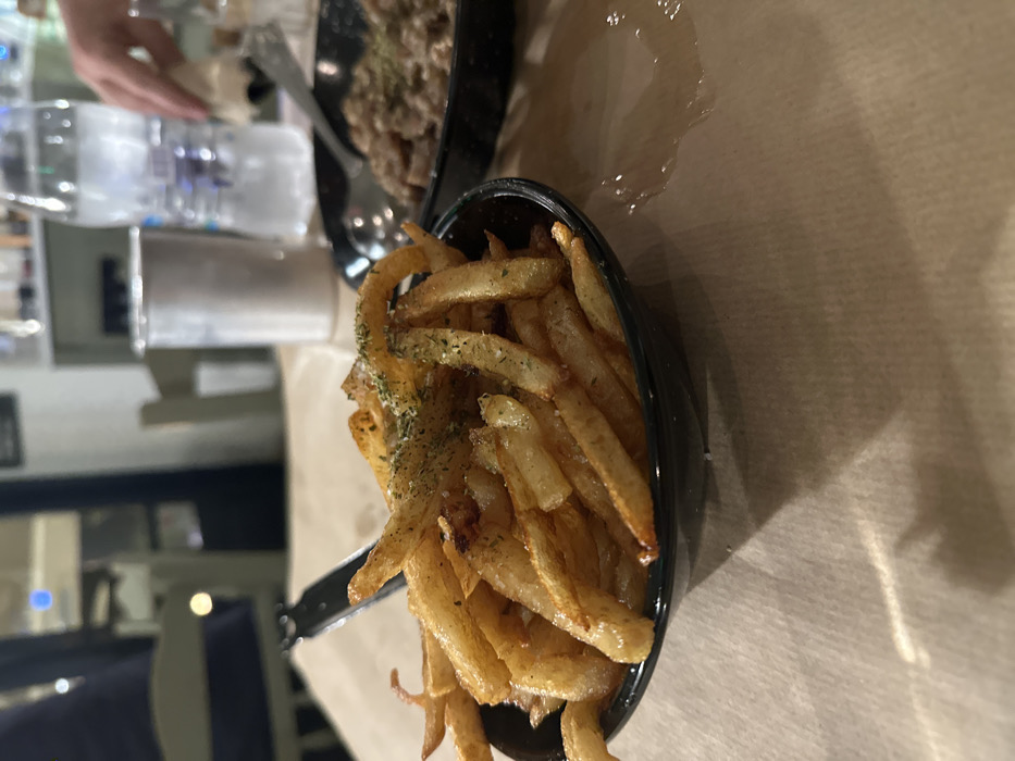 The image presents an angled, close-up view of a dining table, likely in a restaurant or bar setting in Nea Erithrea, Greece. The central subject is a black bowl overflowing with golden-brown french fries, generously seasoned with what appears to be dried green herbs, possibly parsley or dill, and flecks of salt. Some fries exhibit a slight curl, while others are standard straight cuts.
In the blurred background, another dark-colored dish in a black bowl or plate is visible, containing a textured, possibly meat-based or stew-like item, also garnished with herbs. A person's hand is partially visible in the upper left quadrant, reaching towards items on the table. Adjacent to the hand, a clear glass bottle, resembling a water or spirit bottle, stands upright, next to a metallic cylindrical object, possibly a can or container. Cutlery, including a spoon or fork, can be seen near the background dish.
The table surface is covered with a light-brown material, possibly paper or a thin, disposable tablecloth. A noticeable spill or wet mark is visible on this surface, to the right of the bowl of fries. The lighting suggests an indoor setting, likely evening or night. Chairs and blurry background elements hint at a casual dining environment. No discernible text is visible on any objects in the image.