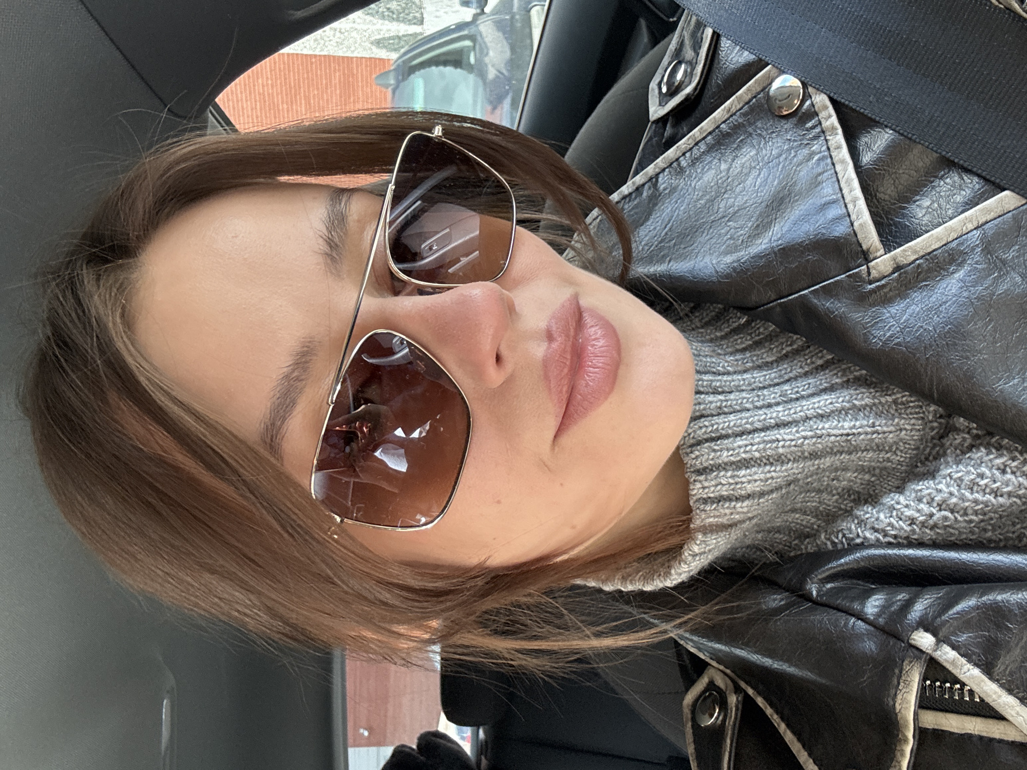 A woman is sitting in a car, wearing sunglasses, a grey knit sweater, and a black leather jacket with white trim. The scene is likely within a car interior. The location in Krasnoyarsk, Russia is not identifiable from the image alone.