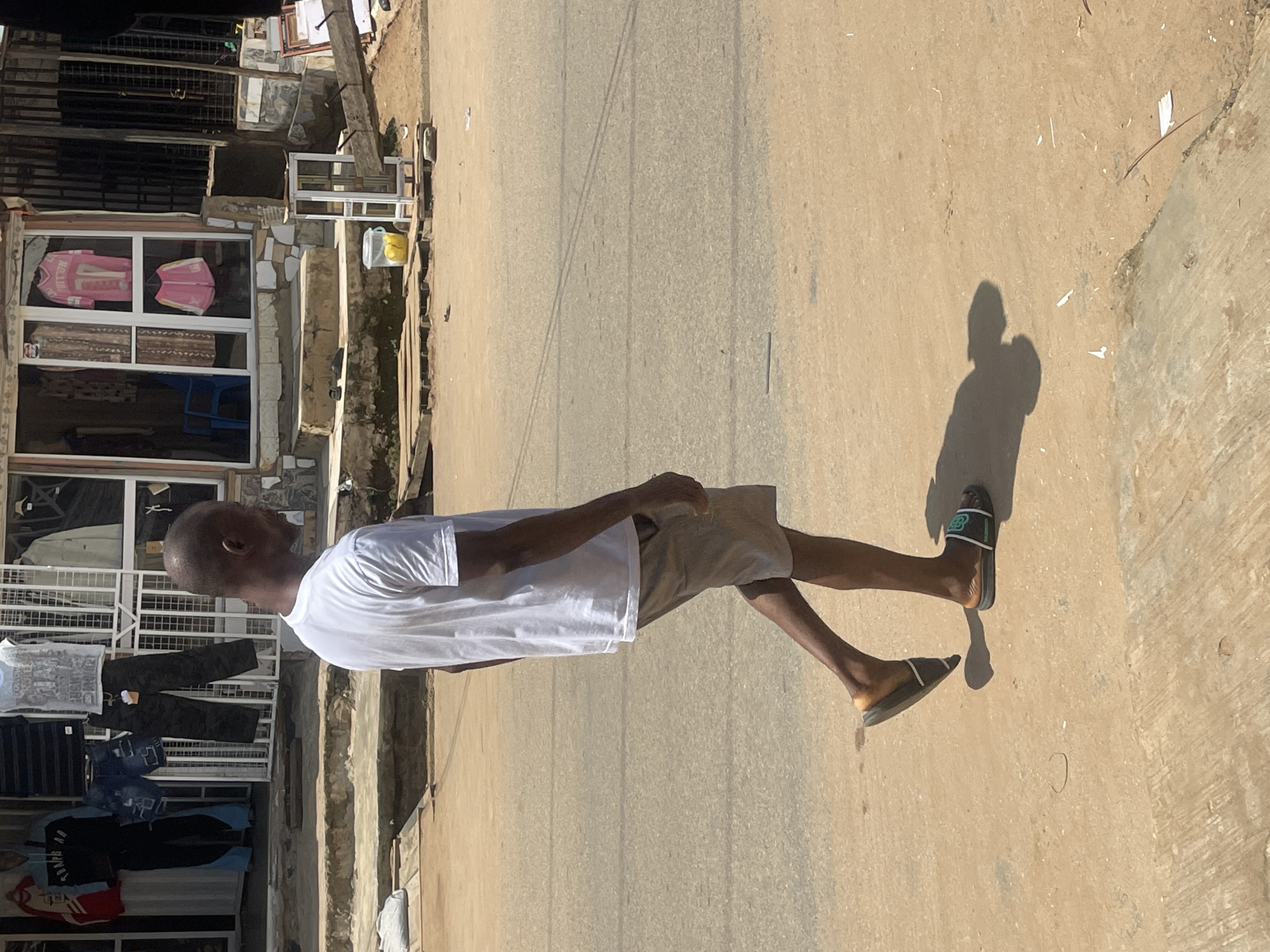 A man wearing a white t-shirt and beige shorts walks on a sandy street in front of a storefront. The storefront has a metal grate covering the display windows, which showcase various clothing items. The man is wearing black and green slides on his feet and casting a shadow on the ground. The environment appears to be a street scene in a city, possibly Abuja, Nigeria, with a road surface made of asphalt and sand. There are some wooden planks and debris on the side of the street. The day is bright and sunny, as indicated by the strong shadows.