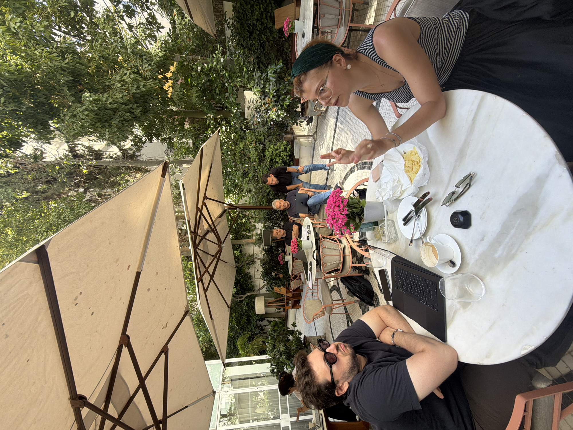 The image depicts an outdoor cafe scene in Tel Aviv, Israel. In the foreground, a man with dark hair, wearing sunglasses and a black t-shirt, sits at a round marble table with his arms crossed. To his right, a woman with blonde hair, wearing a green headband and a striped tank top, is engaged in conversation, gesturing with her hands. On the table between them are a laptop, a coffee cup, a glass of water, a plate of chips, and cutlery.
In the background, several other individuals are seated at tables under large umbrellas. Two men are standing near a table where another man is seated with a laptop. The cafe is surrounded by lush greenery, with trees and plants contributing to a relaxed, open-air atmosphere. The paving of the cafe floor has a patterned design. The overall impression is a casual meeting or work session taking place in an urban outdoor setting.