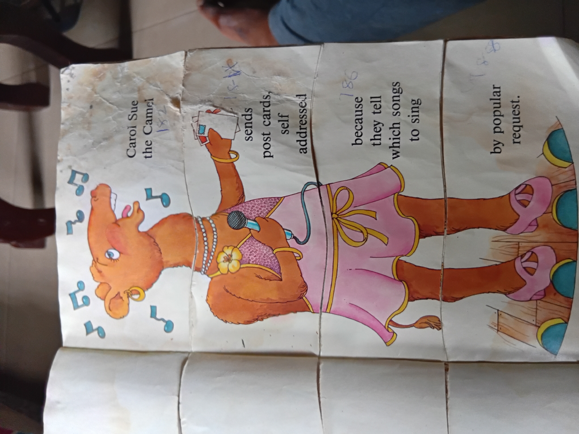 A children's book, open to a page featuring a cartoon illustration of Carol Sue, the Camel. The camel is depicted as a performer, wearing a pink dress with a yellow sash, pearls, a flower accessory, and pink high-heeled shoes. She holds a microphone in one hand and a stack of postcards in the other, with musical notes floating around her head. The text on the page reads: "Carol Sue the Camel 182 sends post cards, self addressed because they tell which songs to sing by popular request." The book is placed on a flat surface, possibly a table, with a person's legs and feet visible in the lower right portion of the image. The environment appears to be indoors, with what looks like a wooden floor. The image was taken in Nkpor, Nigeria.