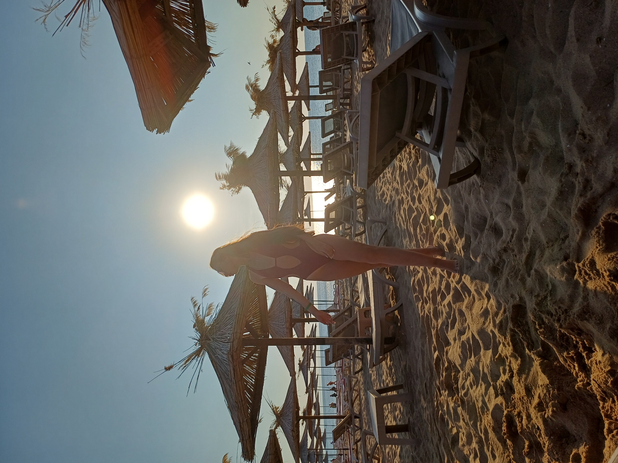 A silhouette of a blonde woman in a one-piece swimsuit stands on a sandy beach in Varna, Bulgaria. She is facing to her left, with her right arm extended. The sun is setting behind her, casting a warm glow across the scene. In the background, there are rows of thatched umbrellas and empty lounge chairs. The sea is visible in the distance. The sand is textured with ripples and shadows.
