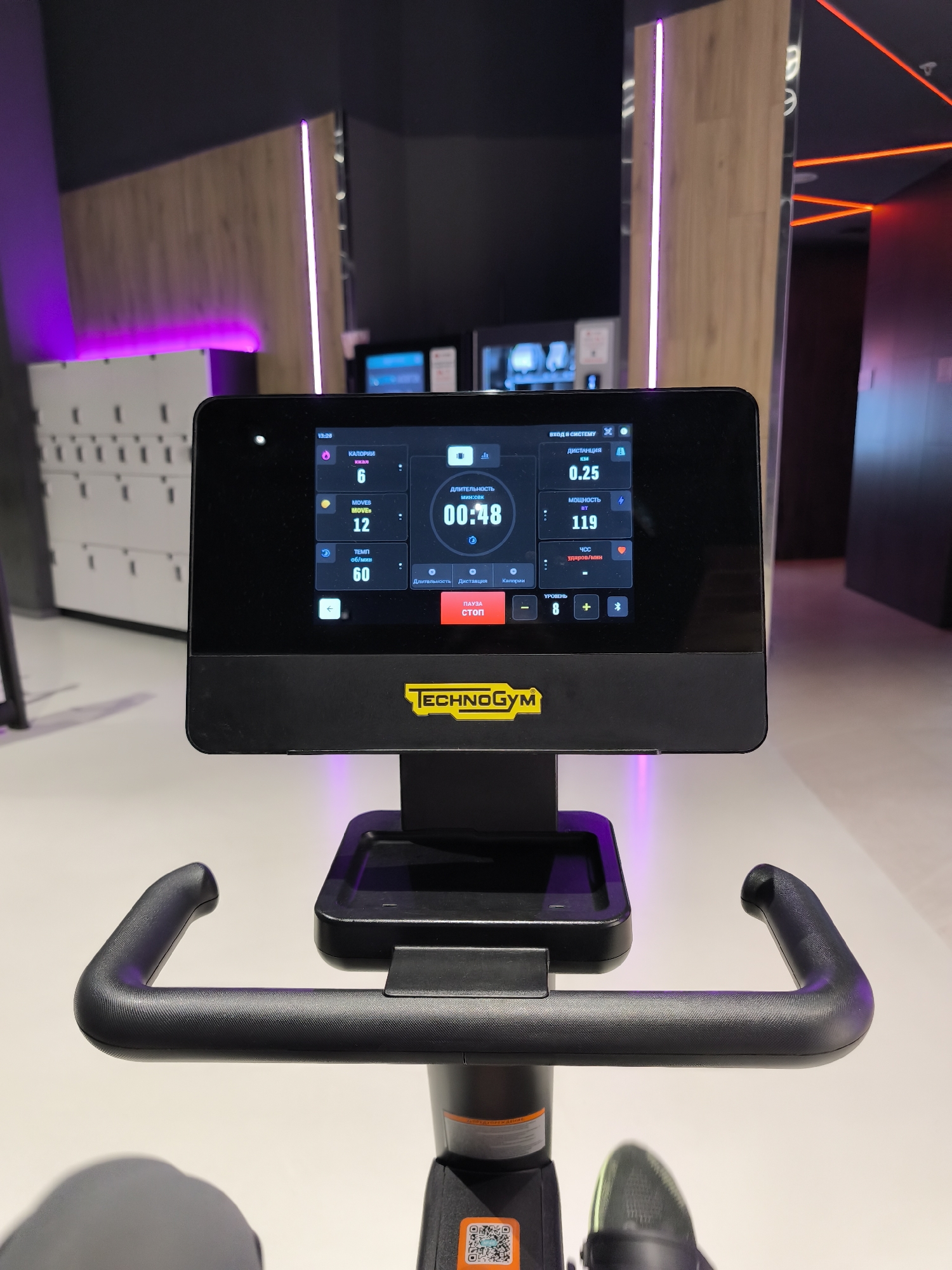 The image shows a Technogym exercise bike in a gym setting. The bike's display screen indicates the following data: 6 calories burned, 12 moves, a tempo of 60 revolutions per minute, a distance of 0.25 kilometers, 119 watts of power, and a heart rate. The timer reads 00:48 (minutes:seconds). The bike's resistance level is set to 8.
The background reveals a modern gym interior. To the left are storage lockers with purple lighting. Through the opening behind the bike, a hallway with what appears to be a lighting installation is visible.
Given the display language (Russian) and the style of the gym, it's likely this image was taken in Russia, possibly Moscow.