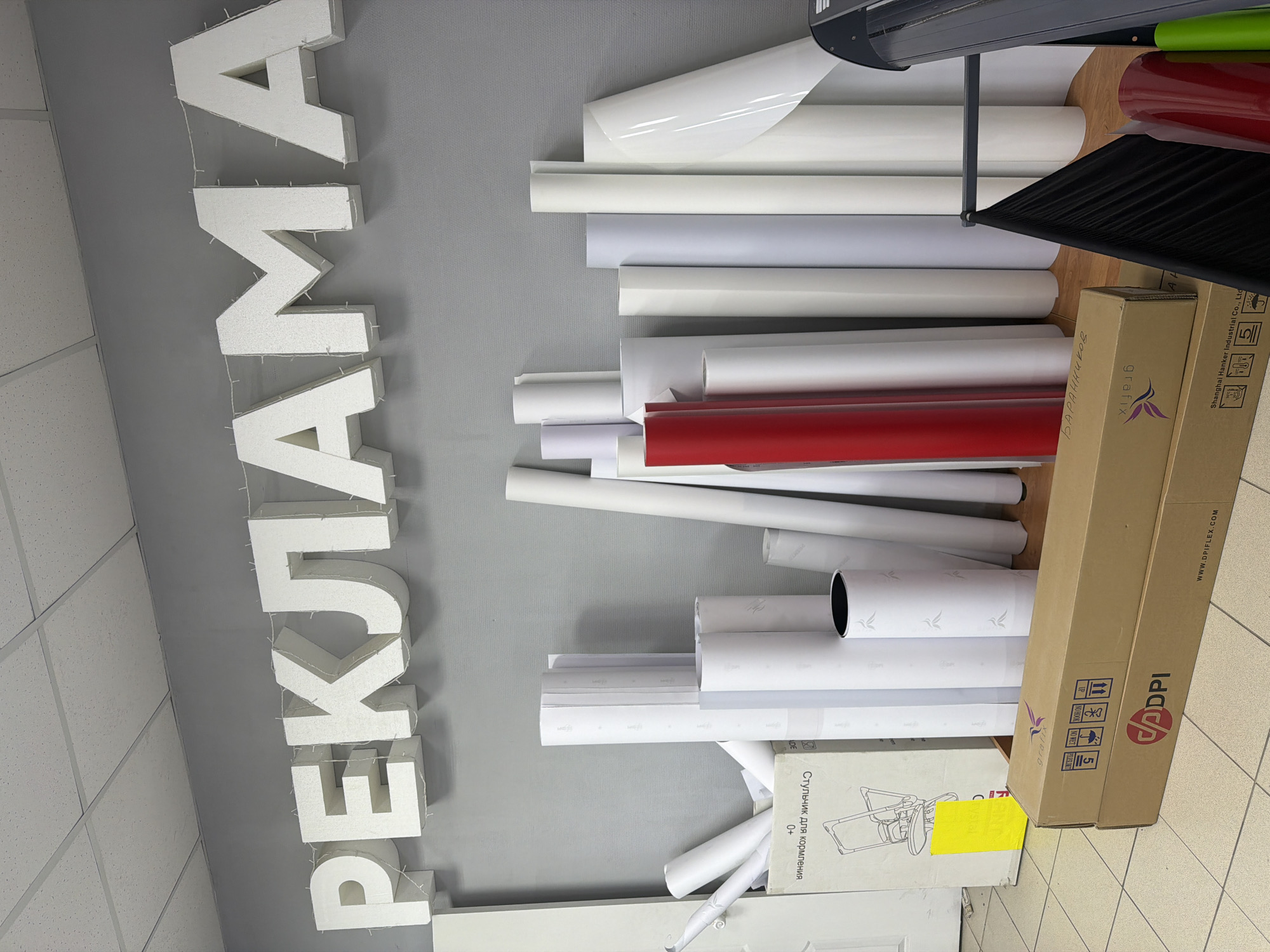 This indoor photograph, taken in Chaikovsky, Russia, captures a cluttered storage or workshop area, likely associated with a sign-making or advertising business. Dominating the left side of the frame are large, white, textured foam letters spelling "РЕКЛАМА" (Advertising) horizontally across a gray wall. These letters are illuminated by small, strung lights.
To the right, numerous rolls of material, predominantly white, with some light gray and a distinct red roll, are stacked vertically and horizontally. These appear to be vinyl or adhesive film rolls, commonly used in sign production. A part of a black, industrial-looking device, possibly a printer or cutter, is visible in the upper right.
Several cardboard boxes are scattered on the floor and stacked among the rolls. One box clearly shows the brand "grafix" and another displays "ADPI" and "WWW.DPIFLEX.COM", along with shipping symbols. Partial text on another box reads "Shanghai Hanker Industrial Co., Ltd". A handwritten name, "Баранников", is visible on one of the boxes. Closer to the bottom, another box is labeled "Стульчик для кормления" (High chair for feeding) with "0+" indicating suitability for infants, suggesting it might be unrelated storage or packaging. The floor is tiled in a light, grid pattern, and the ceiling features standard acoustic tiles. The scene is well-lit by artificial indoor lighting. No people or specific activity are depicted.