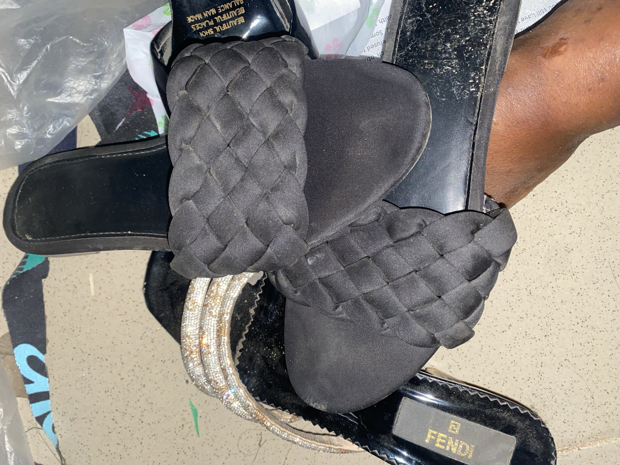 In Jauro Yinu, Nigeria, a close-up shot focuses on a person's foot wearing a black woven slide sandal. Another slide sandal, also black with a woven strap, is positioned in front, partially covering the first sandal. To the left, a bedazzled sandal with multiple rows of rhinestones and a gold trim is visible. The sandals are on a speckled light-colored surface, possibly a floor, with some debris present. Plastic packaging and a torn piece of fabric with blue lettering are in the background.