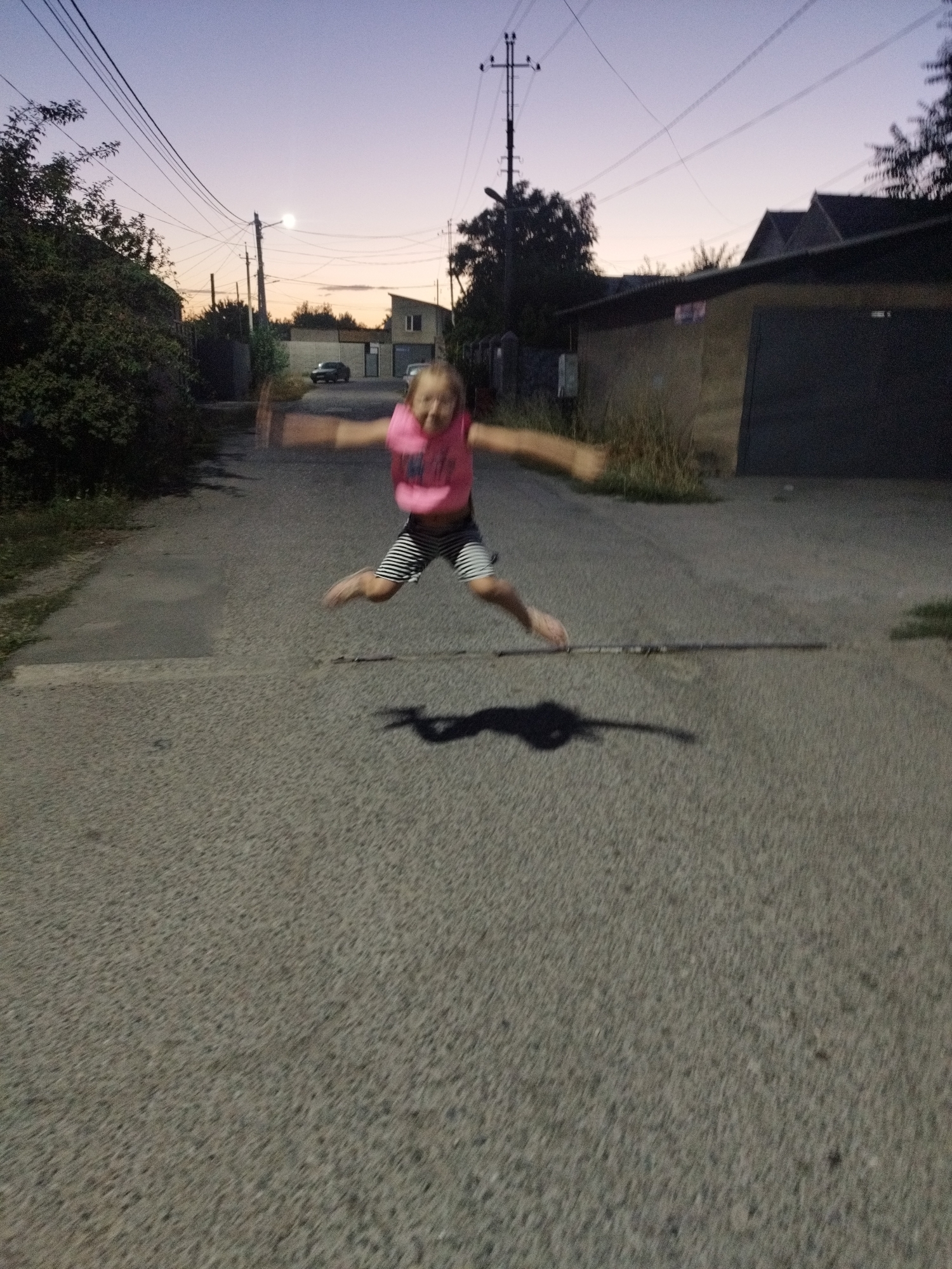 In this image, a young girl in a pink top and striped shorts is captured mid-jump on a paved street during twilight. Her arms are outstretched, and her legs are splayed in the air, creating a dynamic silhouette against the dim lighting. Her face is a blur due to the motion, but her energetic pose suggests a joyful moment. The setting appears to be a residential street, with houses and garages visible on either side. Electrical wires and a utility pole are prominent features in the background, indicating an urban or semi-urban environment. The sky displays a gradient of purple and orange, characteristic of sunset or sunrise. A shadow of the girl is cast on the ground, elongated by the low angle of the light. The overall impression is one of spontaneous childhood exuberance in a quiet, dusky setting.