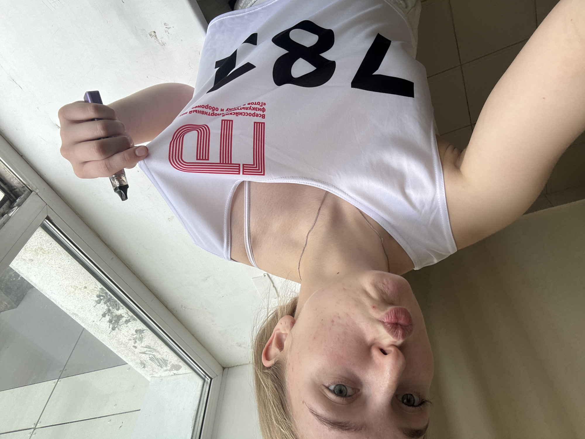 A woman is taking a selfie indoors, possibly in an apartment in Perm, Russia. She is wearing a white tank top with the red letters "ГТО" and the numbers "781" printed on it. The letters stand for "Готов к труду и обороне," which translates to "Ready for Labor and Defense," a Soviet fitness program. She is holding a vape pen in her left hand. The background shows a window with a dirty window sill. The woman's face is visible, with puckered lips.