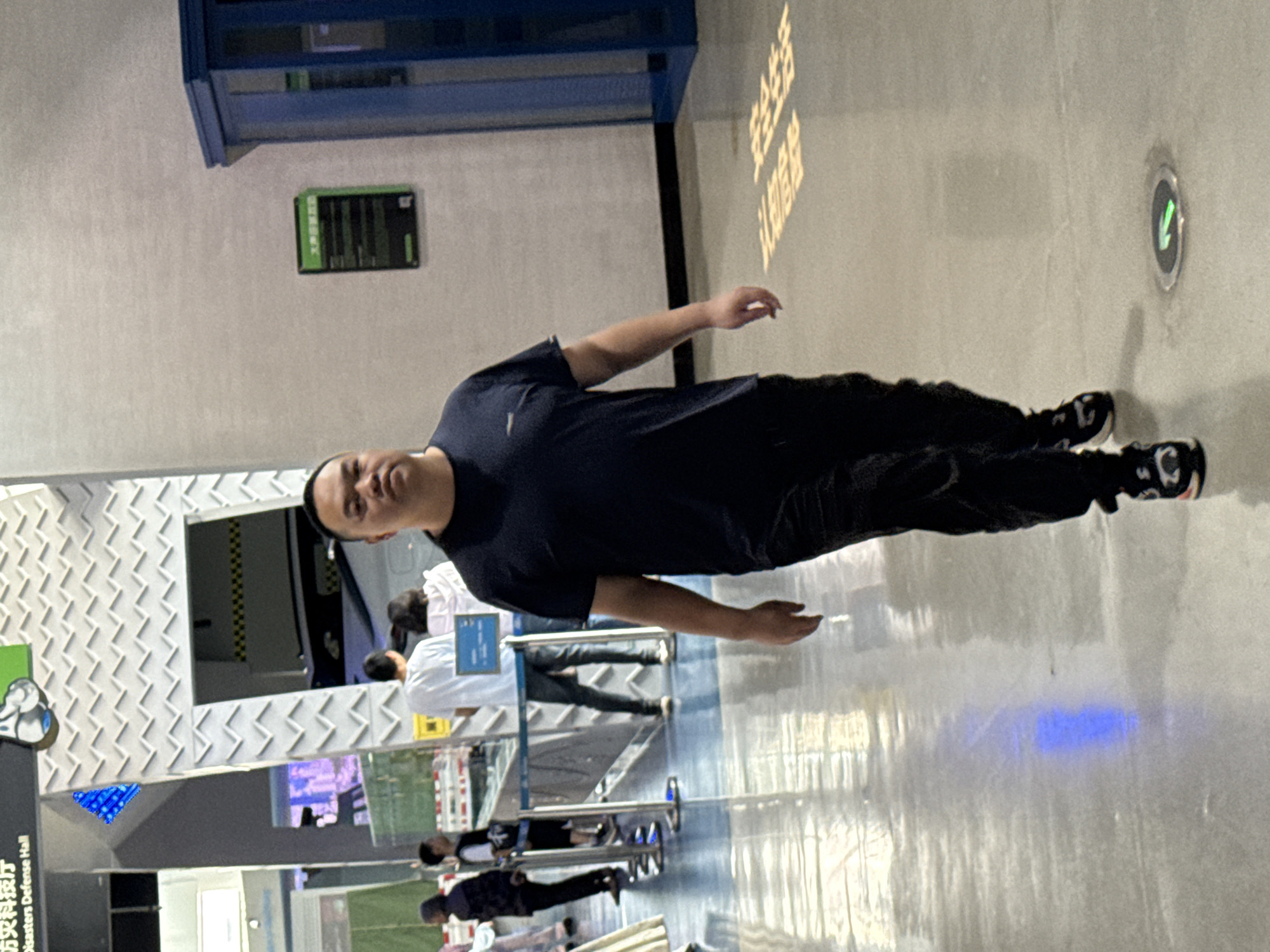 A man wearing a black shirt and black pants stands in what appears to be a modern, indoor space. Other people stand behind him, one wearing a white shirt. The environment is lit with a combination of natural and artificial light. A structure with a diamond-patterned facade is visible to the left. Signage in Chinese indicates that the location is the "Disasters Defense Hall" in the 江北城街道 area of China.