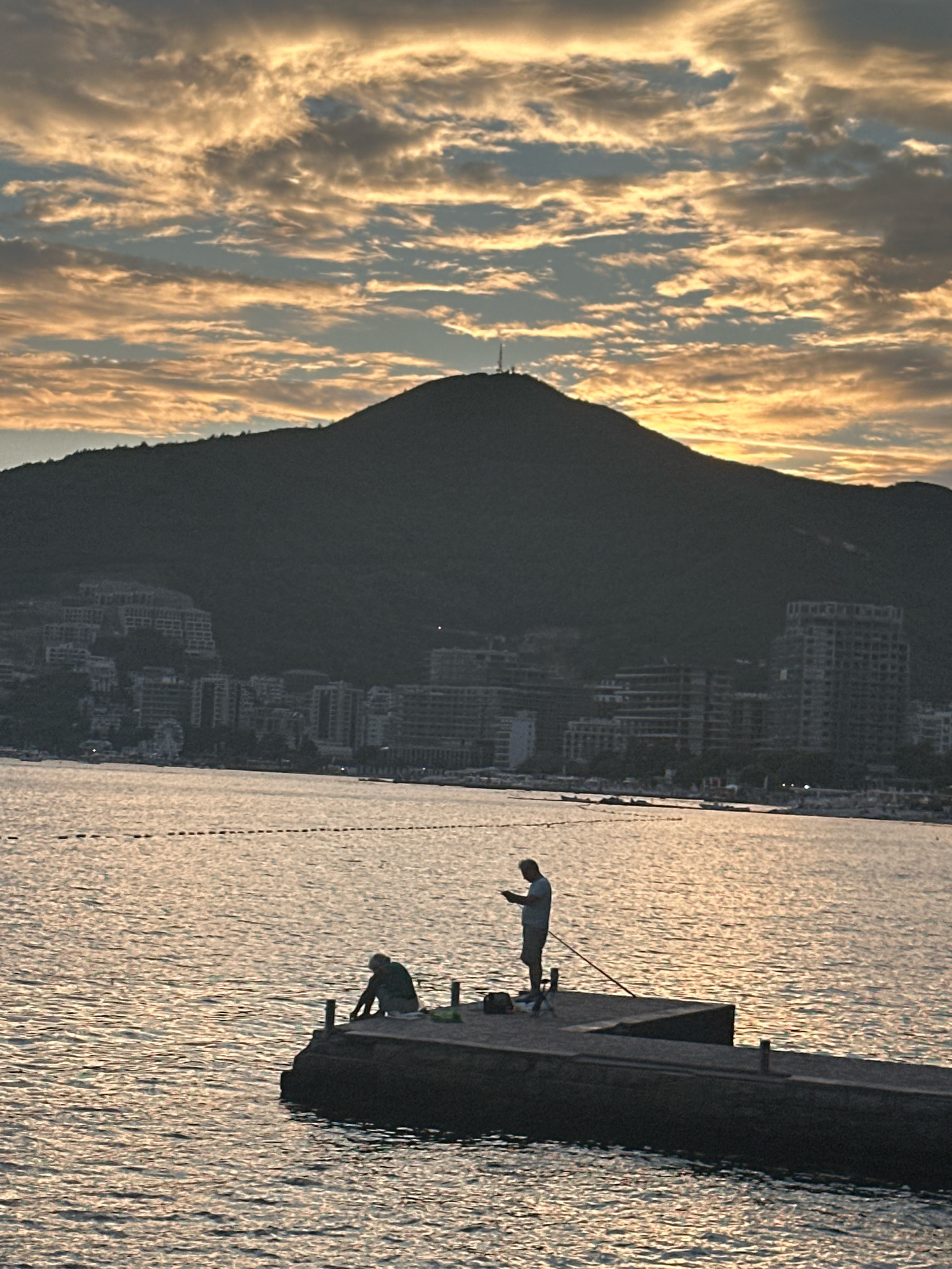 The image depicts a silhouetted view of the city of Budva, Montenegro, at dusk. A large, dark mountain dominates the background, with a small tower visible at its peak. The mid-ground features the Budva skyline, characterized by numerous buildings of varying heights, clustered along the foothills of the mountain. The foreground shows a body of water, likely the Adriatic Sea, with ripples reflecting the golden hues of the setting sun.
On a concrete pier jutting into the water, two individuals are engaged in fishing. One person, in the foreground to the left, is seated on the edge of the pier, facing away from the camera. The second person, standing further along the pier, is holding a fishing rod and looking down at a mobile phone. The water is marked with a series of buoys or markers extending into the distance. The sky is filled with dramatic clouds tinged with yellow and orange, indicative of a sunset. The overall atmosphere is serene and peaceful.