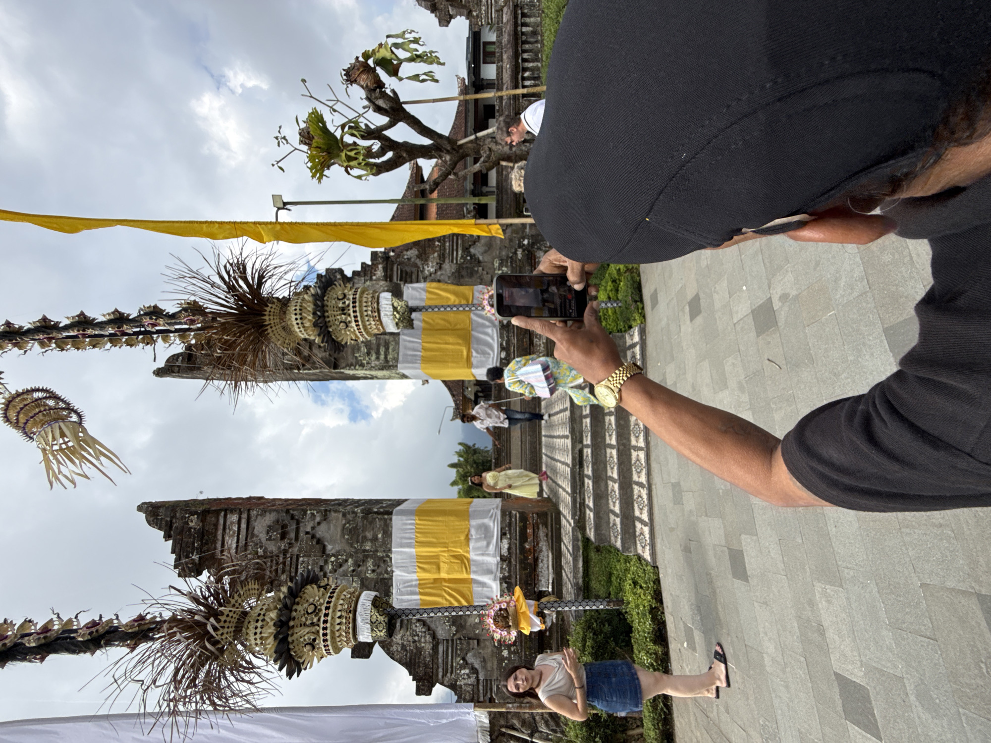 In the city of Baturiti, Indonesia, a person wearing a black cap and black shirt is seen from behind, holding up a smartphone to capture an image. The phone is pointed towards a traditional Balinese temple gate, characterized by its stone structure and intricate carvings. Two large white and yellow banners hang prominently on either side of the gate. Above the gate, decorative elements made of dried leaves and woven materials are suspended, along with a long yellow flag. In front of the gate, several people are visible. On the left, a woman in a denim skirt and a light-colored top stands on a paved area. Further back, a man and a woman in colorful traditional attire are walking up the temple steps. The ground in front of the temple is paved with large gray stone tiles. The sky is overcast with visible clouds. The overall scene captures a moment of cultural tourism at a significant architectural landmark.