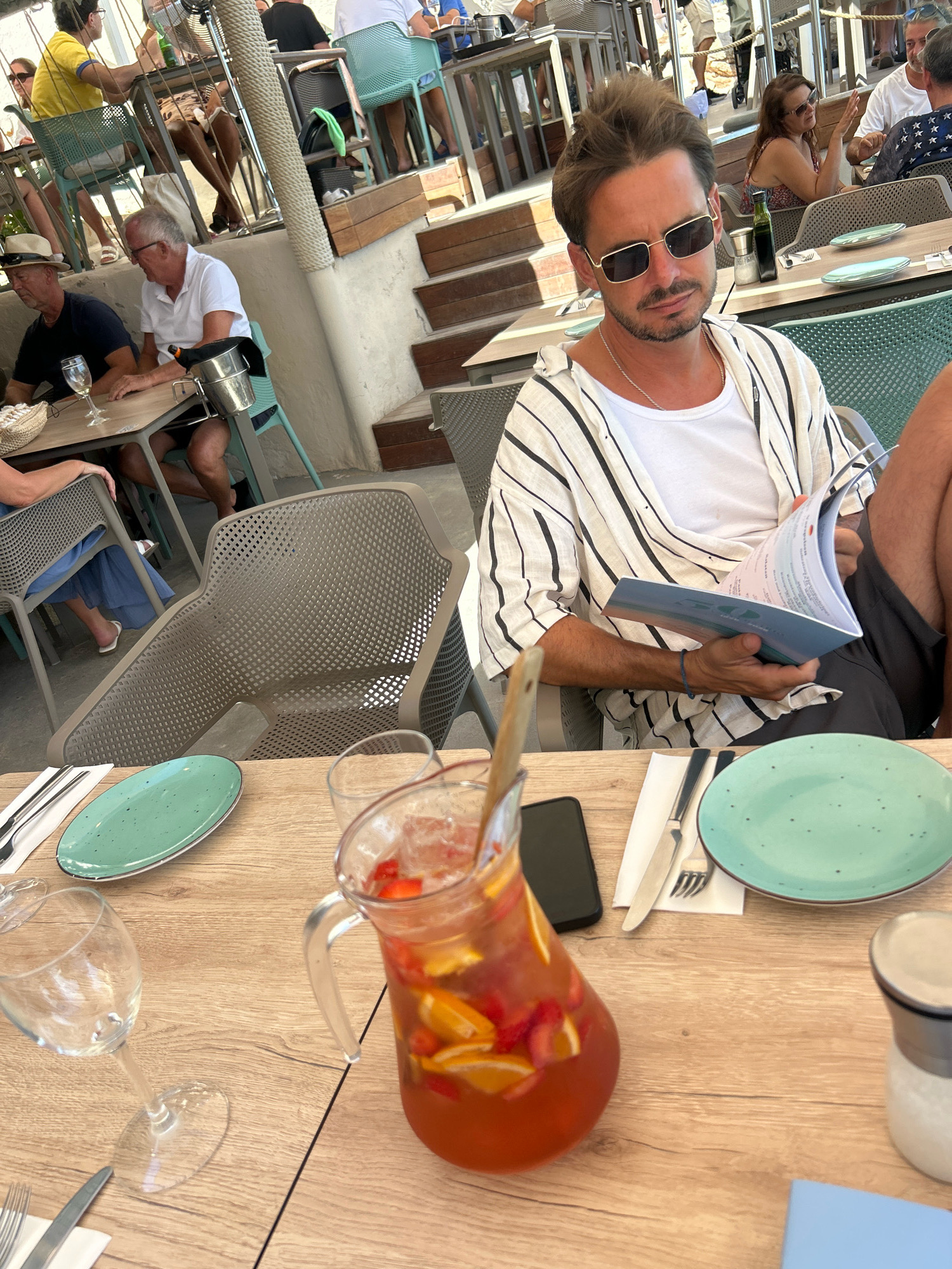 The image depicts an outdoor dining setting at a restaurant in Cas Català-ses Illetes, Spain. The scene is centered on a table with various items, including a pitcher of sangria filled with fruit, a wine glass, plates, cutlery, and a salt shaker. In the foreground, a man wearing a white and black striped shirt over a white t-shirt and sunglasses is seated, holding a menu. He has short brown hair and a beard. Behind him, other patrons are visible, some seated at tables and others walking in the background. The environment suggests a sunny day with a relaxed atmosphere, typical of a Mediterranean coastal resort. The architecture and decor, including outdoor seating and umbrellas, further reinforce the location.